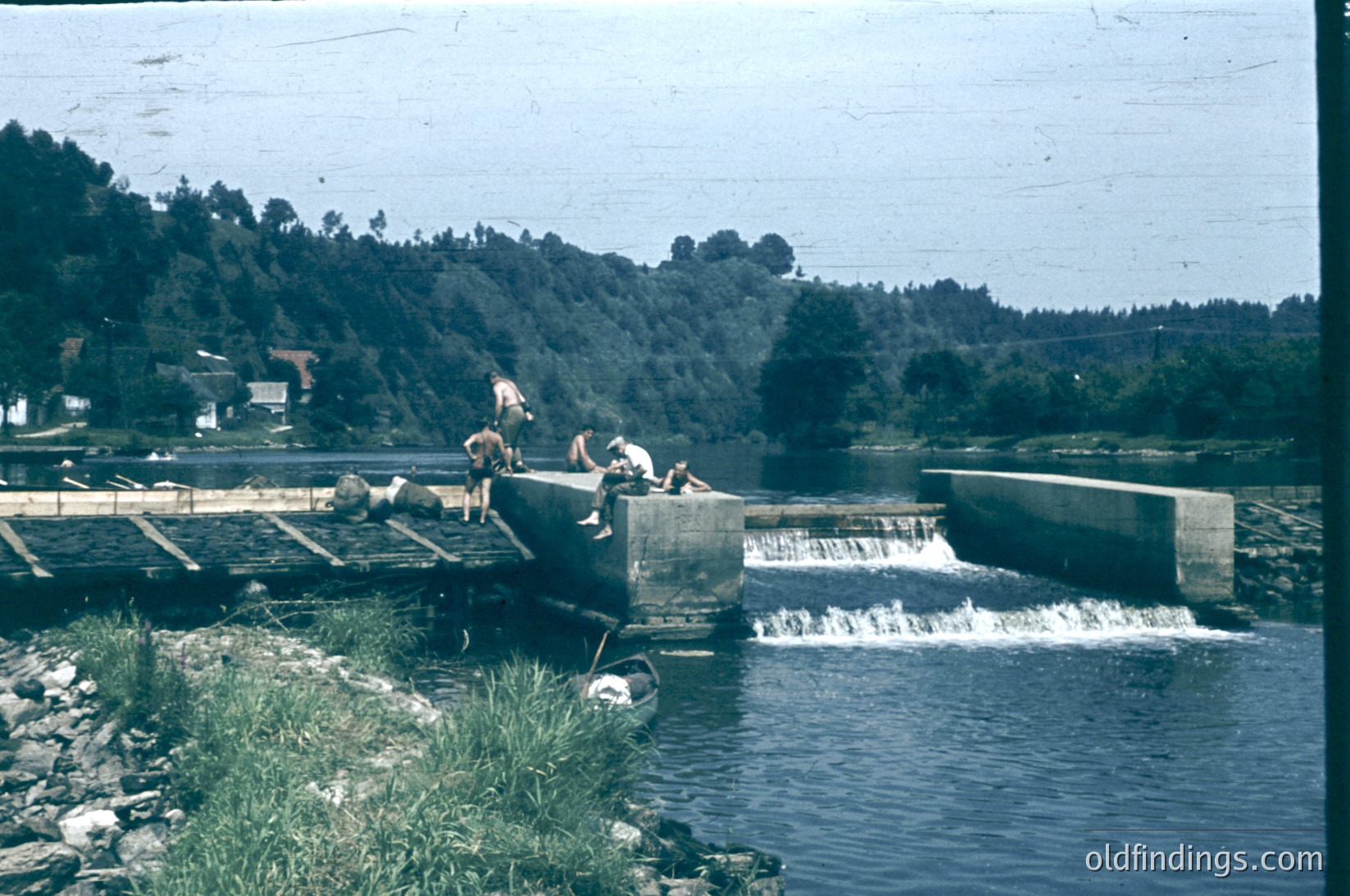 Vintage black-and-white photo of three men repairing a concrete dam spillway, mid-20th century. Rural landscape with forested hills and a small village in background. Industrial infrastructure meets natural scenery.