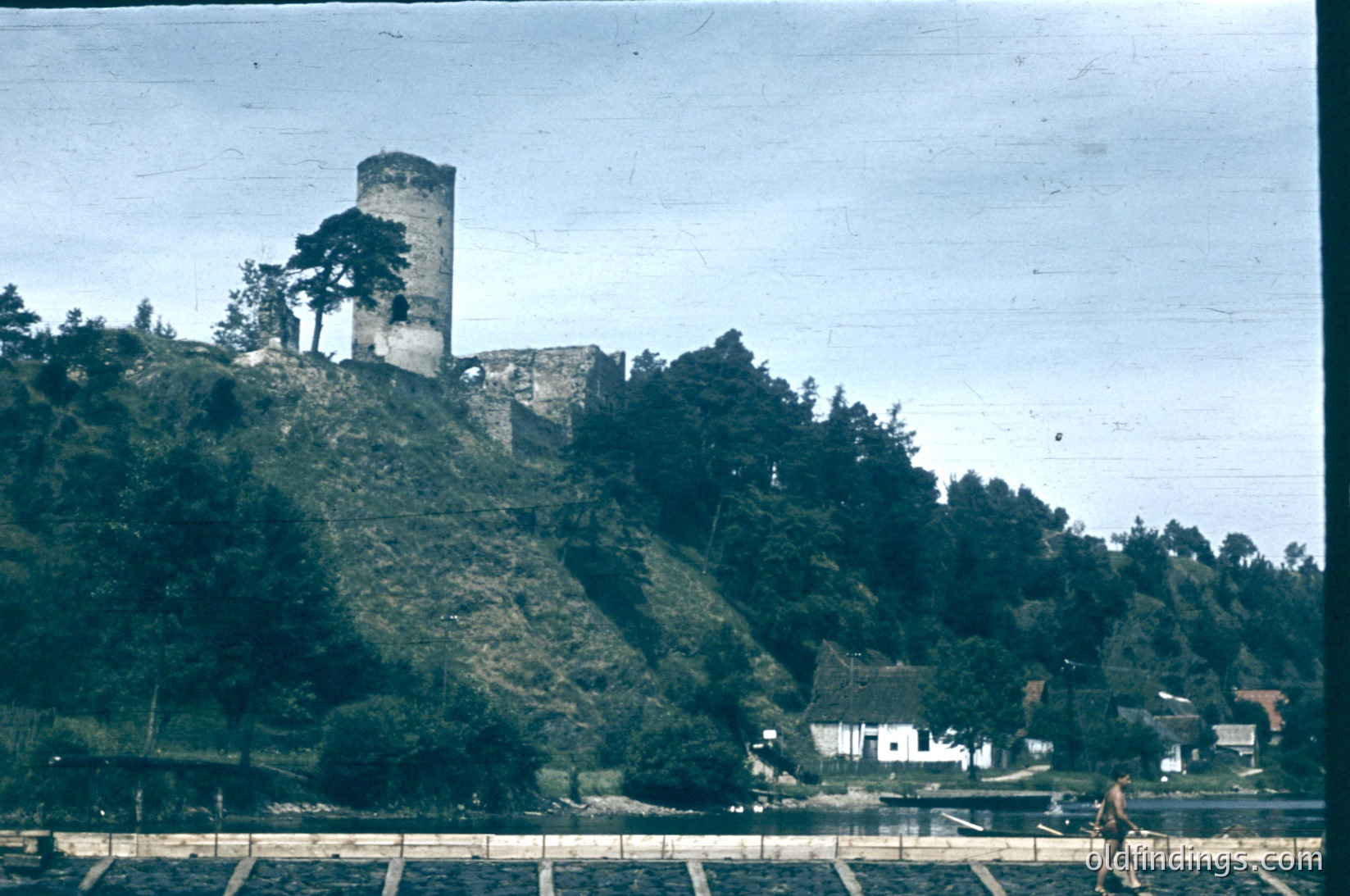 Vintage sepia-toned photo of a medieval stone tower perched atop a forested hillside, overlooking a serene lake. Remnants of a larger castle complex are visible. A lone figure walks along a stone path near the water’s edge, suggesting a quiet, rural setting. Likely Eastern European, mid-20th century.
