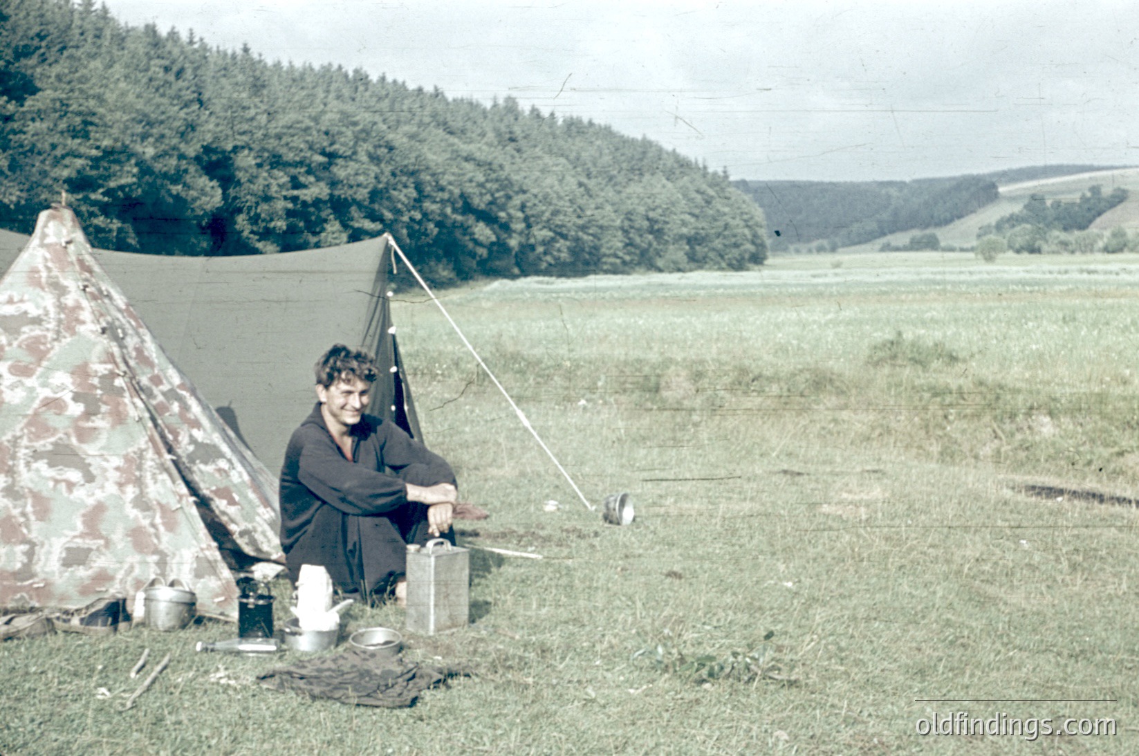 Vintage camping scene in a rural, forested landscape with rolling hills. A person sits on a folded blanket beside a camouflage-patterned tent, preparing food on a portable stove. Mid-20th century outdoor lifestyle, likely 1950s–1960s.