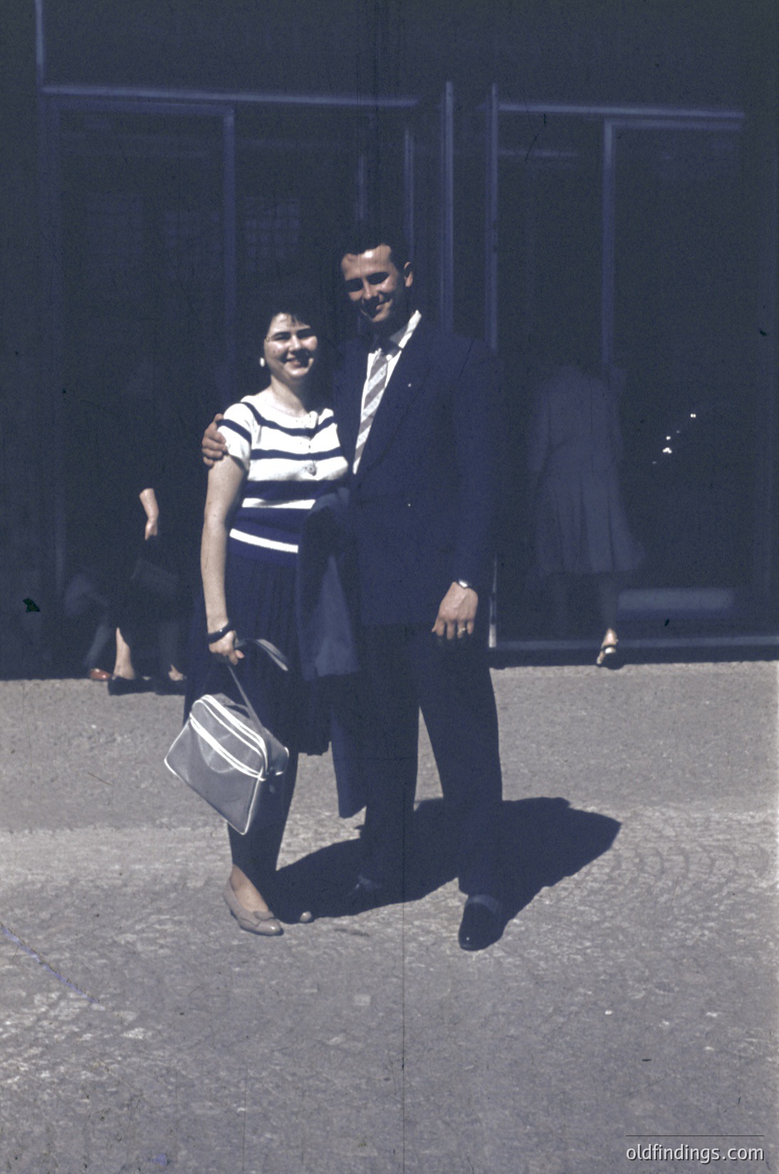Couple posing outdoors in 1960s-era attire—man in dark suit with white shirt, woman in striped dress with sandals and suitcase. Urban setting with concrete pavement and industrial-style building. Warm sepia tone suggests vintage photography.