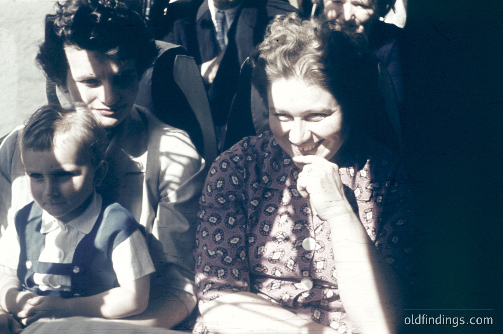 Vintage sepia-toned photo of a family group in mid-20th century attire, likely 1940s–1950s. A woman in patterned blouse and apron holds a child in a striped shirt, while a man in a dark jacket stands behind them. Crowd of indistinct figures in the background suggests public or communal setting. Warm sepia tones enhance nostalgic, historical feel.