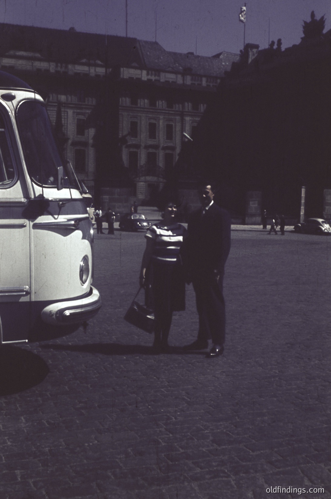 Mid-20th century urban scene featuring a vintage white taxi (likely a Type 2) parked on cobblestone streets. A man in a dark suit and hat stands beside the vehicle, holding a briefcase. Historic European architecture with classical facades and a flagpole in the background suggests a European capital.