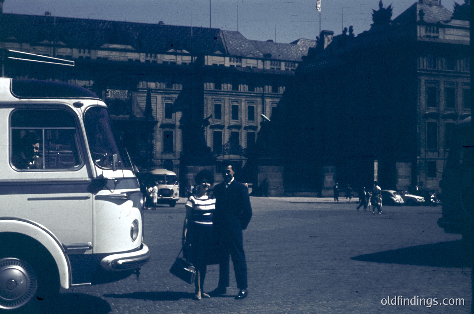 Vintage urban scene featuring a man and woman in mid-20th-century attire (1950s-60s) standing near a vintage bus in a European city square. Historic architecture with ornate facades and cobblestone streets visible. Warm sepia-toned filter enhances nostalgic atmosphere.