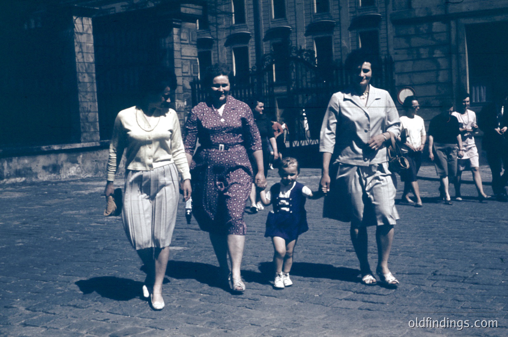 A family of four walks through a European city street in the 1950s, holding hands. The woman in the center wears a floral dress with a belt, flanked by a woman in a striped dress and a man in a light blazer. The child wears a striped shirt and shorts. Cobblestone streets and stone buildings with arched doorways frame the scene.