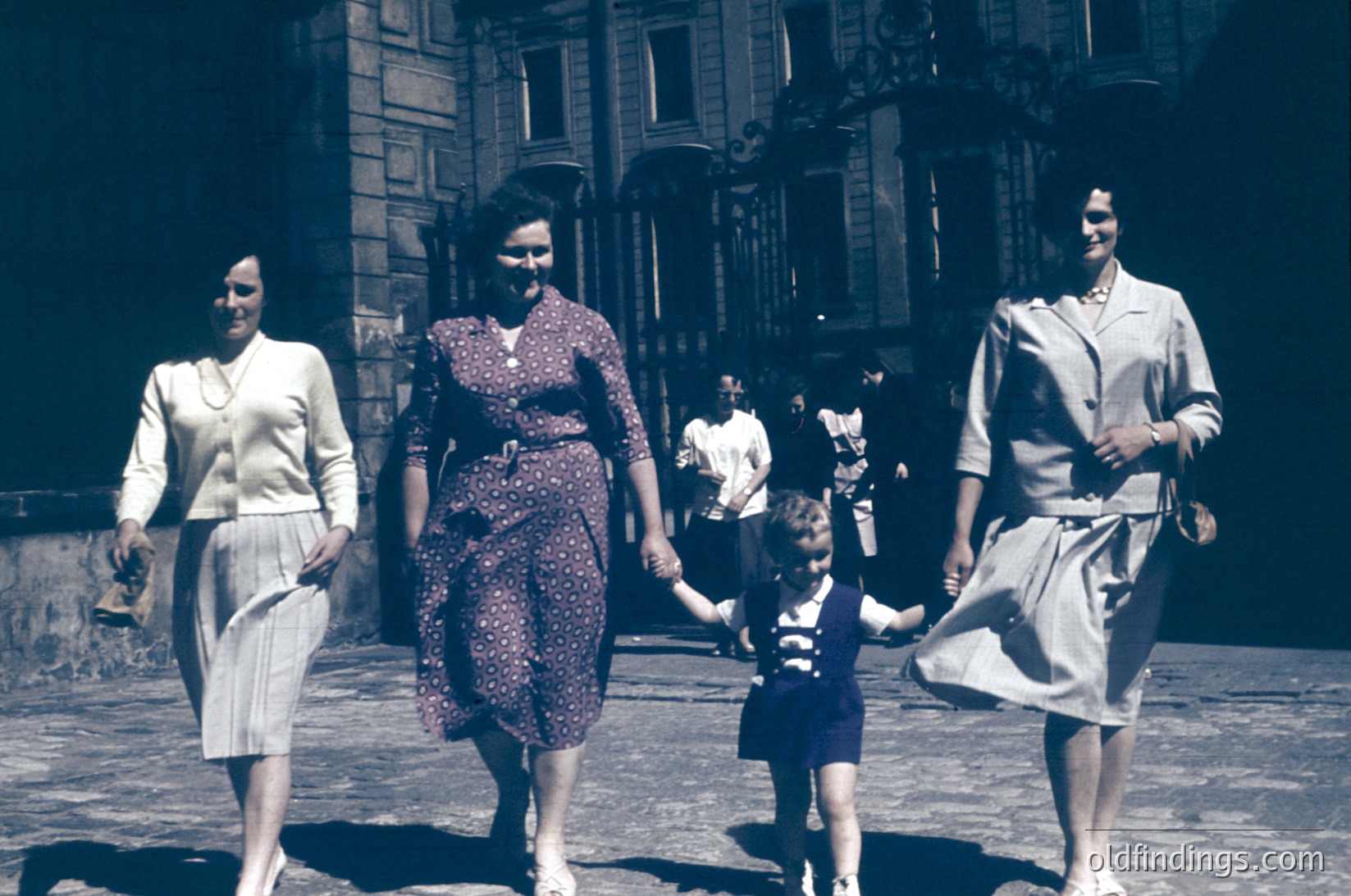Three women and a child walk hand-in-hand on a European street, 1950s. Architectural details include ornate balconies and wrought-iron railings. Women wear structured suits and polka-dot dresses; child wears a striped shirt and shorts. Sepia-toned vintage photograph.