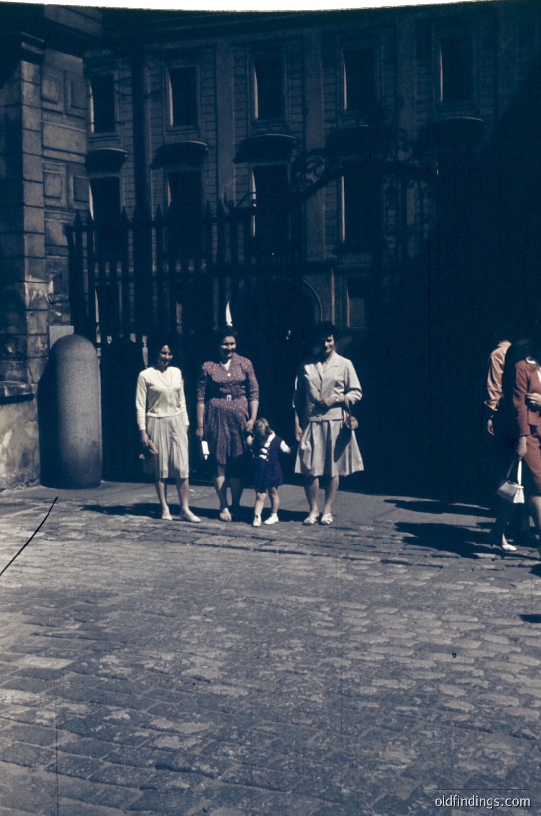 Vintage sepia-toned street scene featuring four women and a child in mid-20th-century attire, likely 1950s–1960s. Women wear knee-length dresses and headscarves; one holds a child’s hand. Cobblestone pavement and stone arches with cylindrical columns frame the scene. Urban European architecture suggests a historic district.