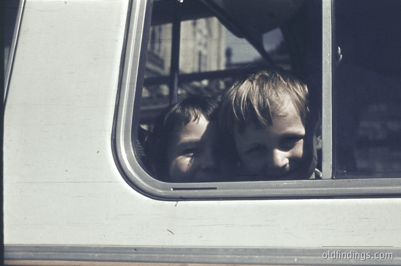 Two children peer out of a vintage bus window, framed by the metal sill. Their expressions suggest curiosity or contemplation. The bus interior shows worn vinyl seats and a partial view of the driver’s area. Likely mid-20th century, possibly or , in a Western or European setting.
