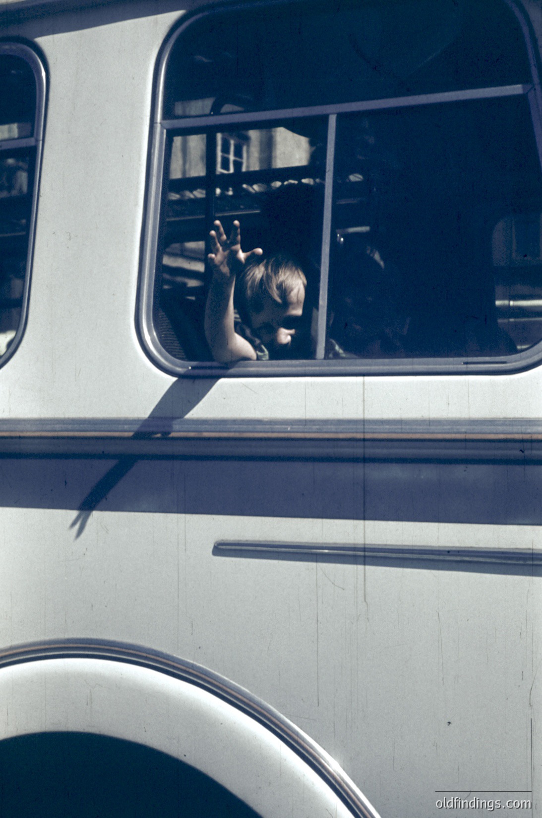 Child waving from a vintage double-decker bus interior, likely mid-20th century. Reflective windows and metal handrails visible. Urban setting suggested by bus design and lighting.