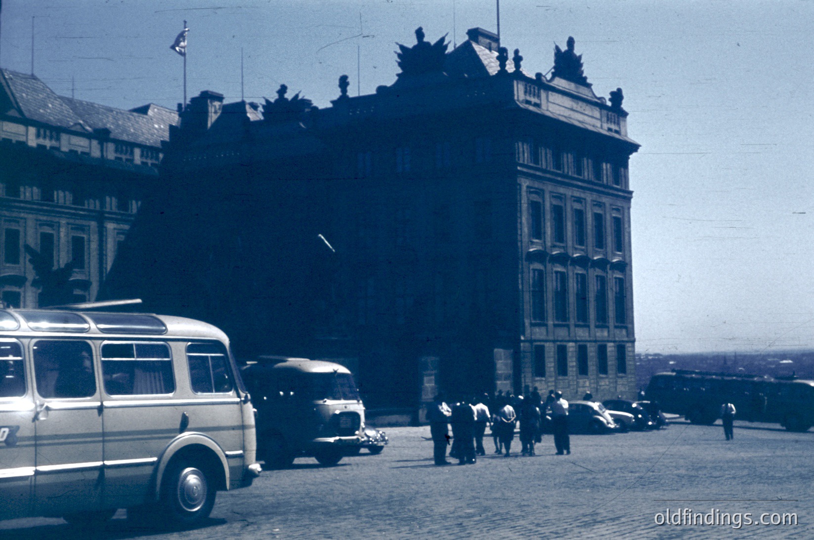 Vintage black-and-white street scene featuring a grand, ornate building with classical architecture—crenellated roofline, arched windows, and decorative stonework. Mid-20th century vehicles (bus, vintage cars) and pedestrians populate a cobblestone plaza. Likely European urban setting, possibly .
