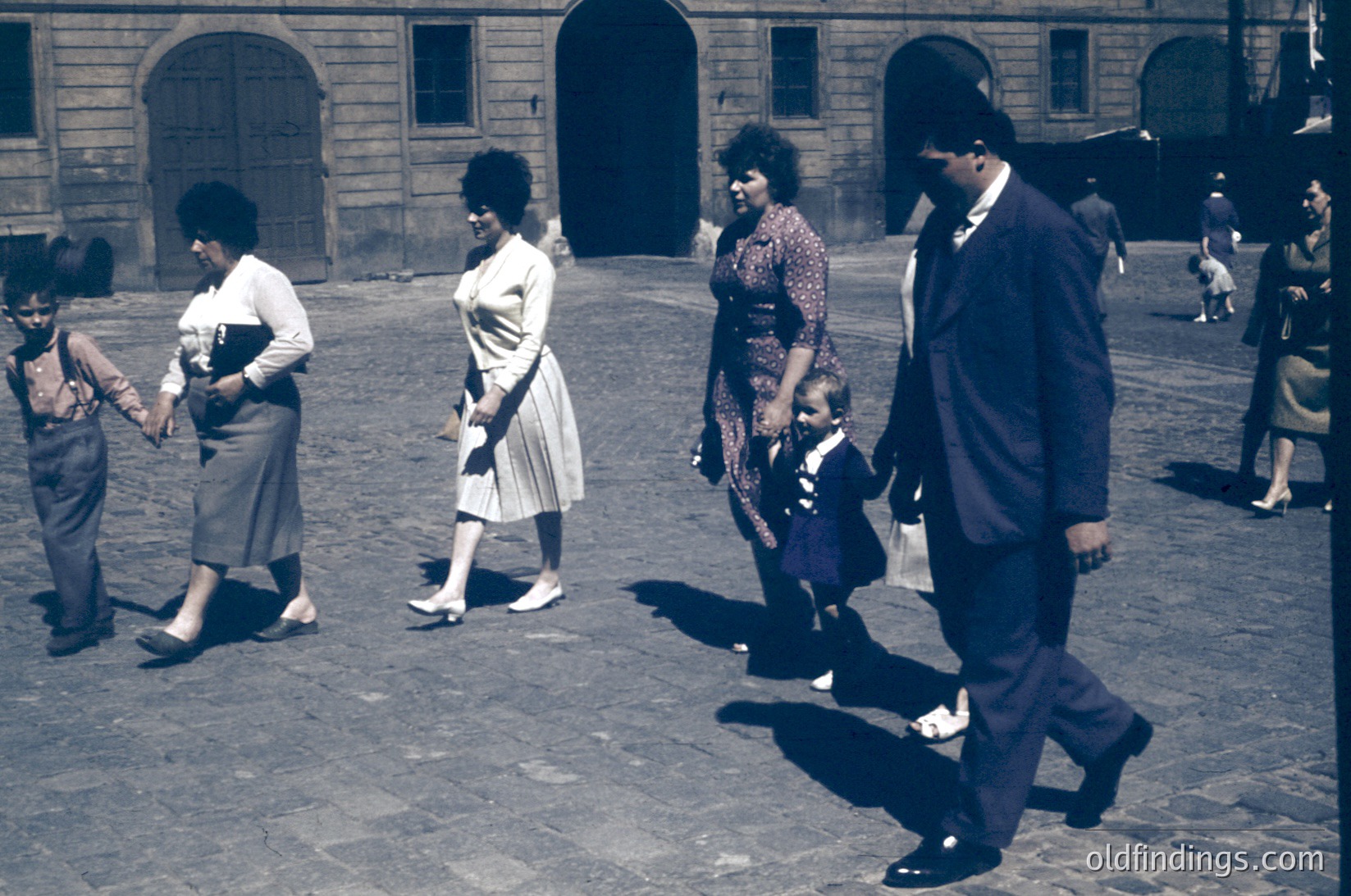 Vintage sepia-toned street scene featuring a family of five walking in an urban courtyard. Women wear 1960s-style dresses with pleats; men in suits and overalls. Stone archways and cobblestone ground suggest European architecture. Likely mid-20th century.