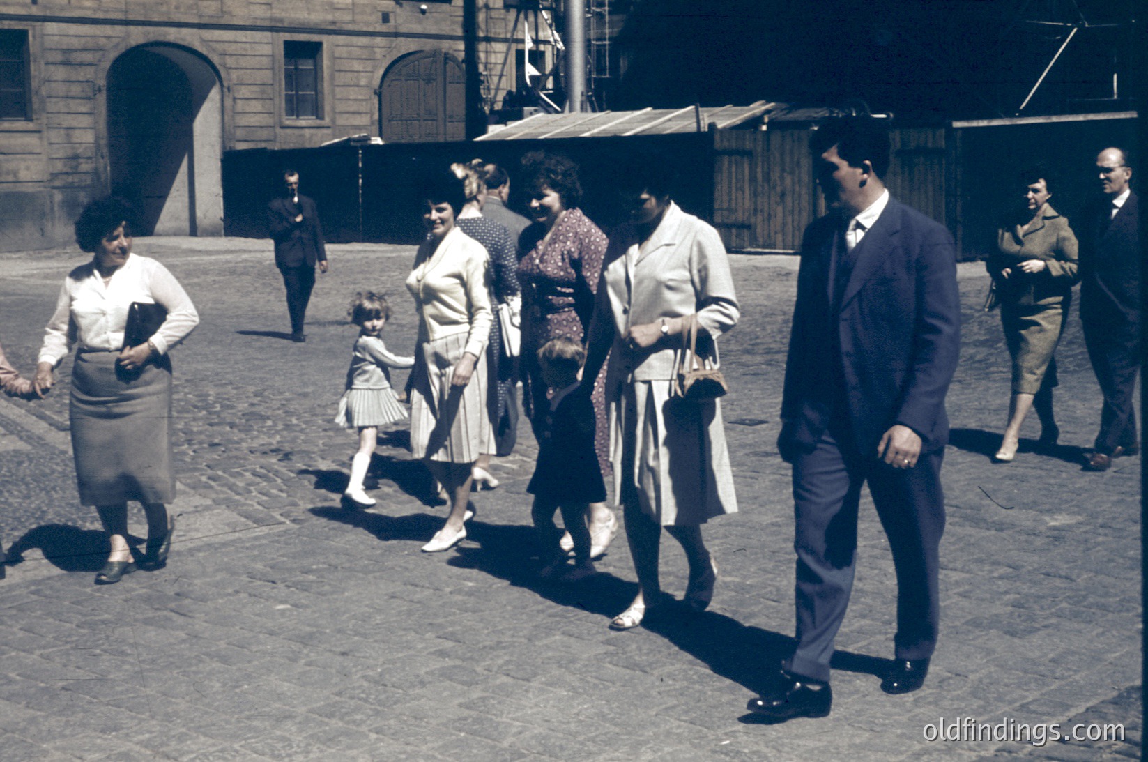 Vintage street scene featuring mid-20th century attire—women in full skirts, cardigans, and headscarves; men in suits and ties. Cobblestone pavement and industrial brick buildings suggest an urban European setting. Likely 1950s–1960s era.