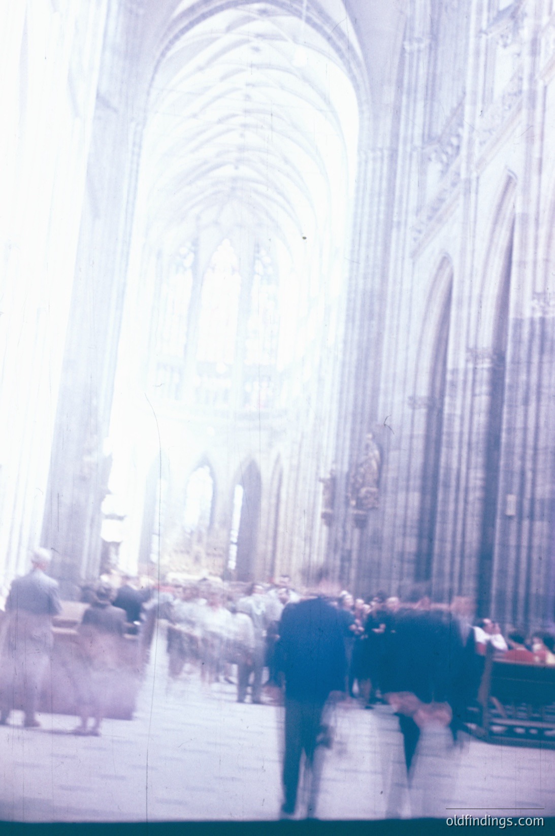 Vintage photograph of Gothic-style cathedral interior with blurred motion of mid-20th-century visitors. Tall, ribbed vaults and pointed arches dominate the space. Light filters through stained-glass windows, casting soft hues. Likely European, 1950s-1960s.