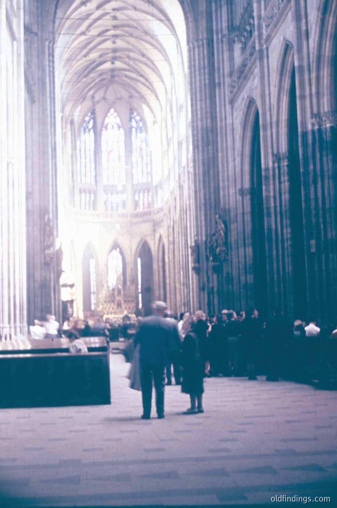 Gothic cathedral nave with pointed arches, stained-glass windows, and stone columns. Blurred figures in 1960s-era clothing (men in suits, women in dresses) suggest mid-century European setting. Warm light filters through stained glass, illuminating the stone floor. Ideal for historical architecture studies or vintage travel inspiration.