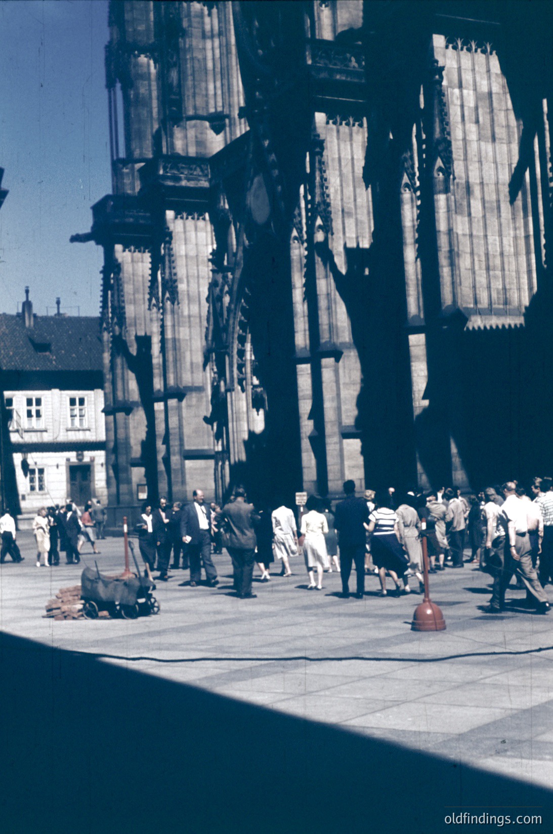 Mid-20th century urban scene featuring Gothic-style scaffolding on a grand cathedral façade. Crowd of casually dressed individuals—men in suits, women in dresses—walking and standing on a paved plaza. Red fire hydrant and scattered debris on ground. Likely post-WWII restoration work in a European city.