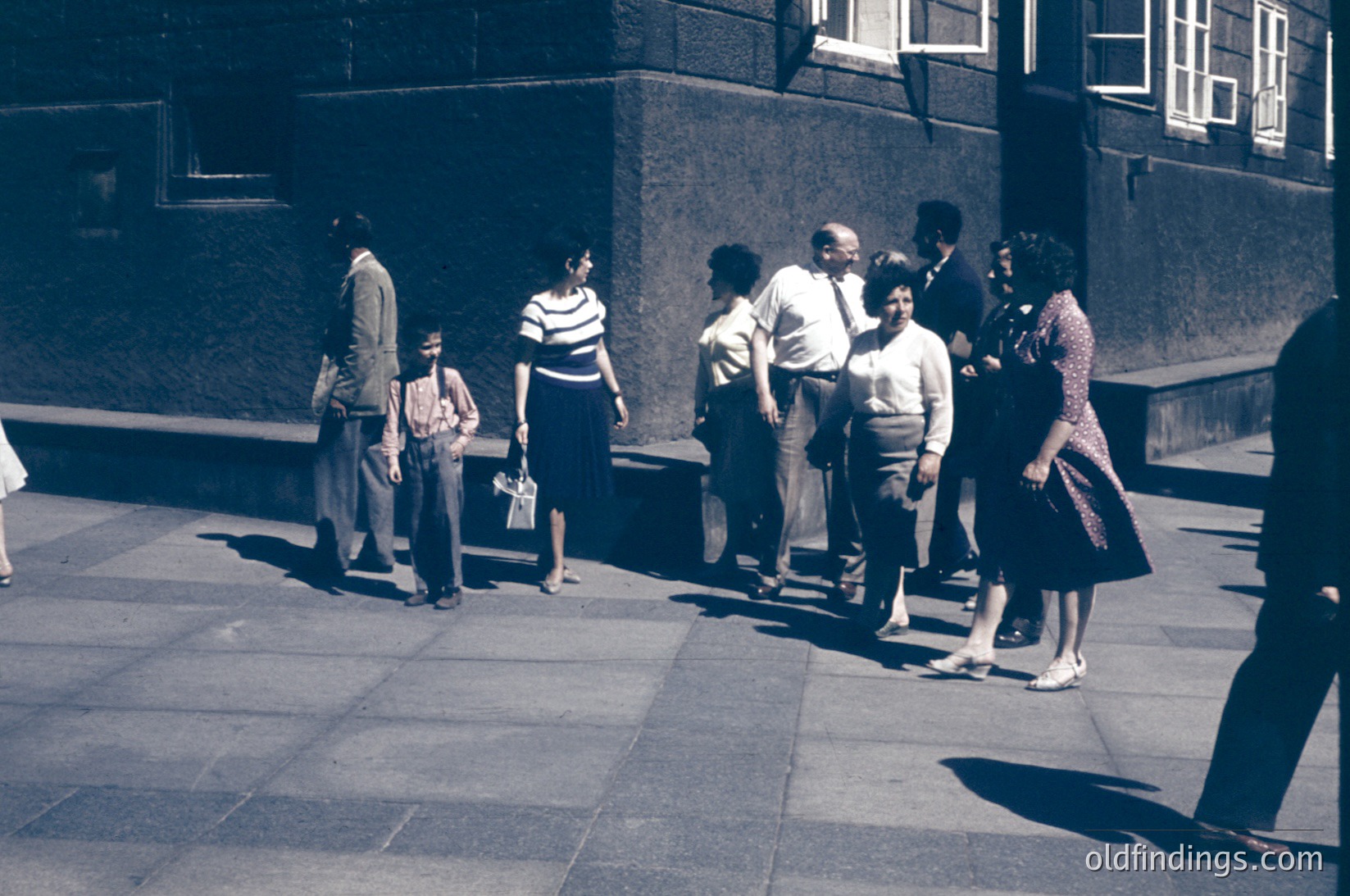 Group of 10+ people in 1950s-60s streetwear—men in suits, women in knee-length dresses and cardigans—gathering near a mid-century concrete building. Urban setting with geometric shadows on pavement.