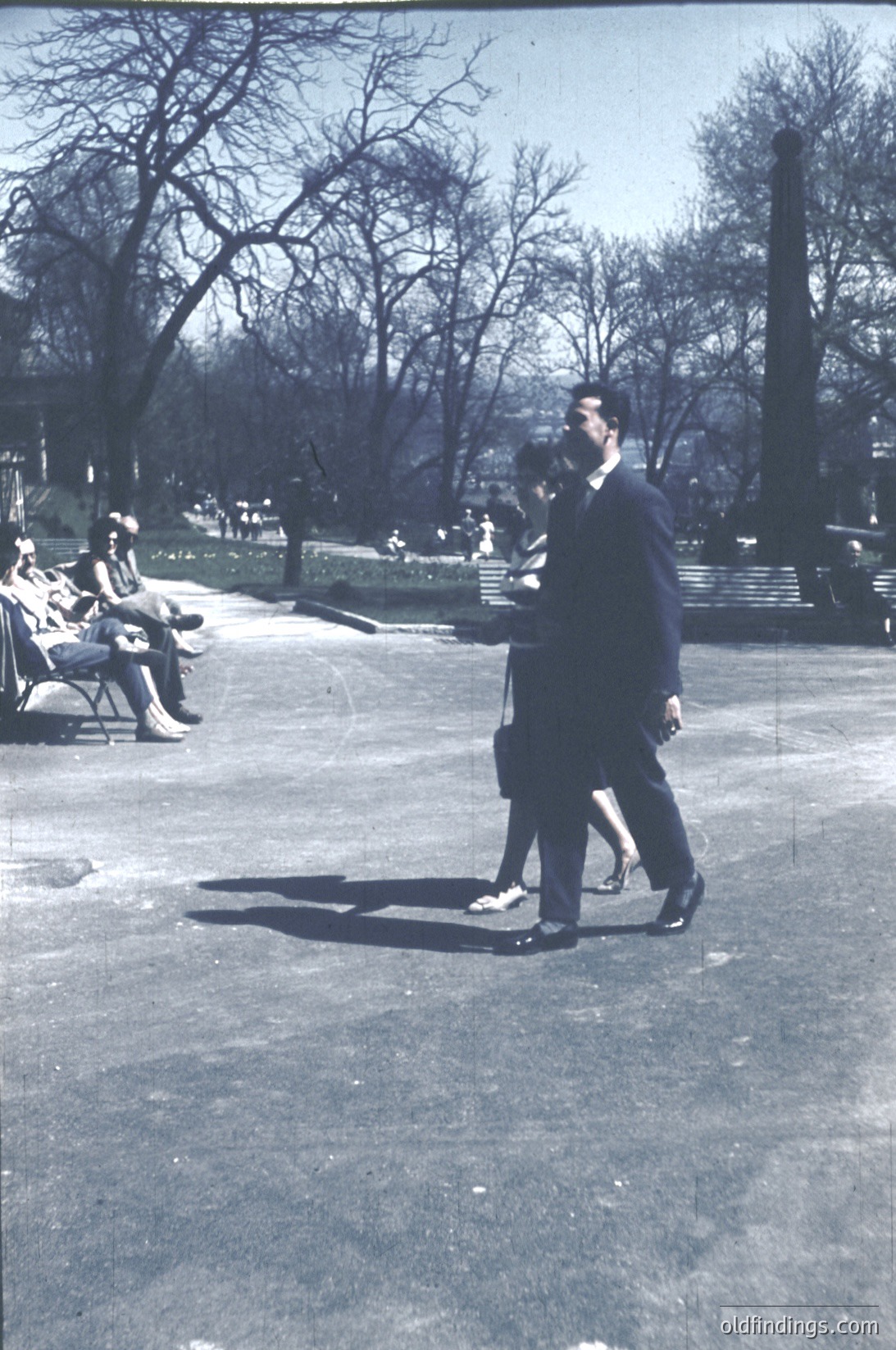 Black-and-white photo of two men in formal attire (dark suits, ties) walking in a park. One man holds a cigarette. Leisurely parkgoers sit on benches in the background. Bare trees suggest late winter/early spring. Urban park setting, likely mid-20th century.