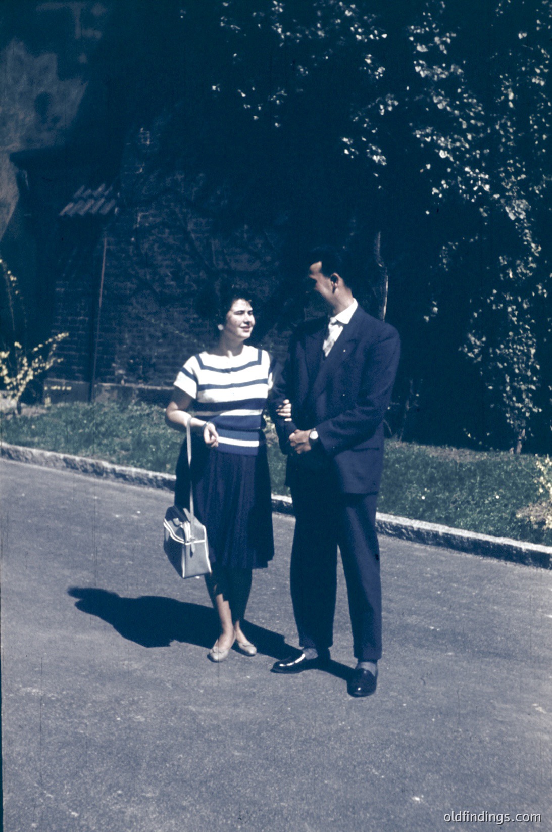 Couple in mid-20th century attire—woman in striped blouse & knee-length skirt, man in suit—posing outdoors on paved path. Lush greenery and trees in background suggest park or garden setting. Handheld camera case hints at leisurely outing. Likely or , Western or East Asian influence.