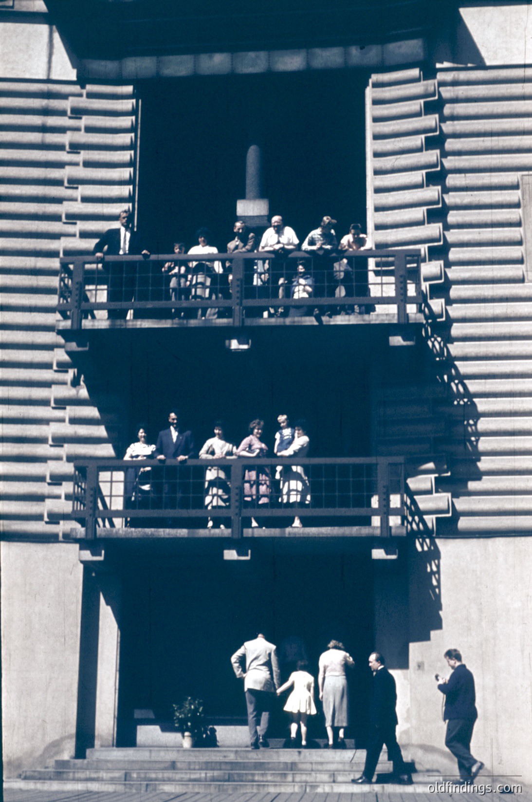 Black-and-white photo of a multi-level outdoor staircase gathering, likely 1950s–1960s. Two groups: one seated on upper balcony-style platform, another descending stairs. Formal attire suggests a social or ceremonial event. Architectural details include concrete steps and metal railings.