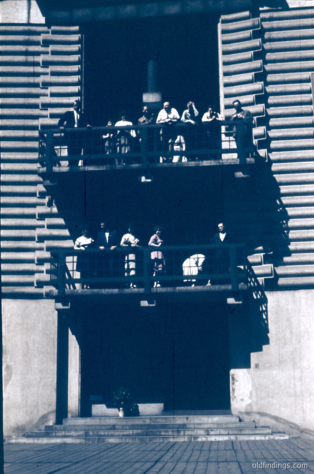 Vintage black-and-white photo of a multi-level outdoor seating area with wooden slatted railings, likely a public terrace or viewing platform. Groups of people sit on benches, engaged in conversation or relaxation. The architectural style suggests a mid-century modern or Brutalist influence. The setting appears urban, with a tiled ground surface and minimalist design.