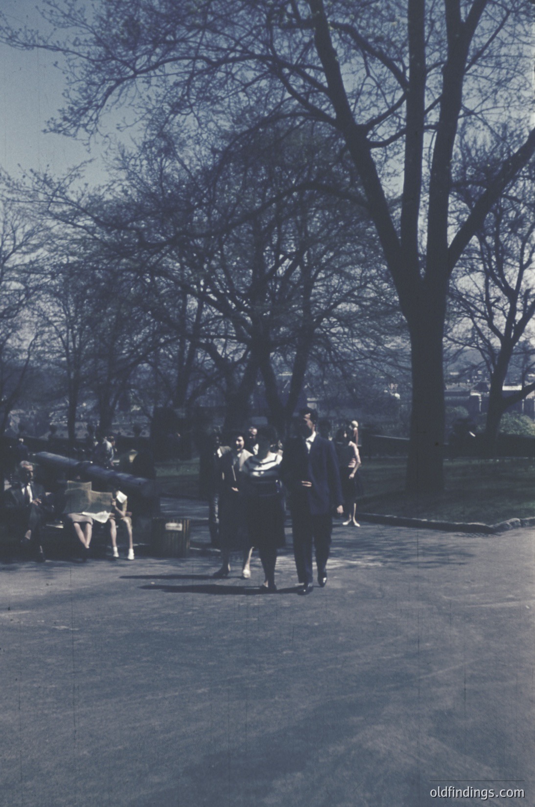 Vintage sepia-toned park scene with 1950s-60s fashion: group of men in suits and women in dresses walking near a bench. Leaves suggest autumn. Urban park setting with trees and distant buildings.