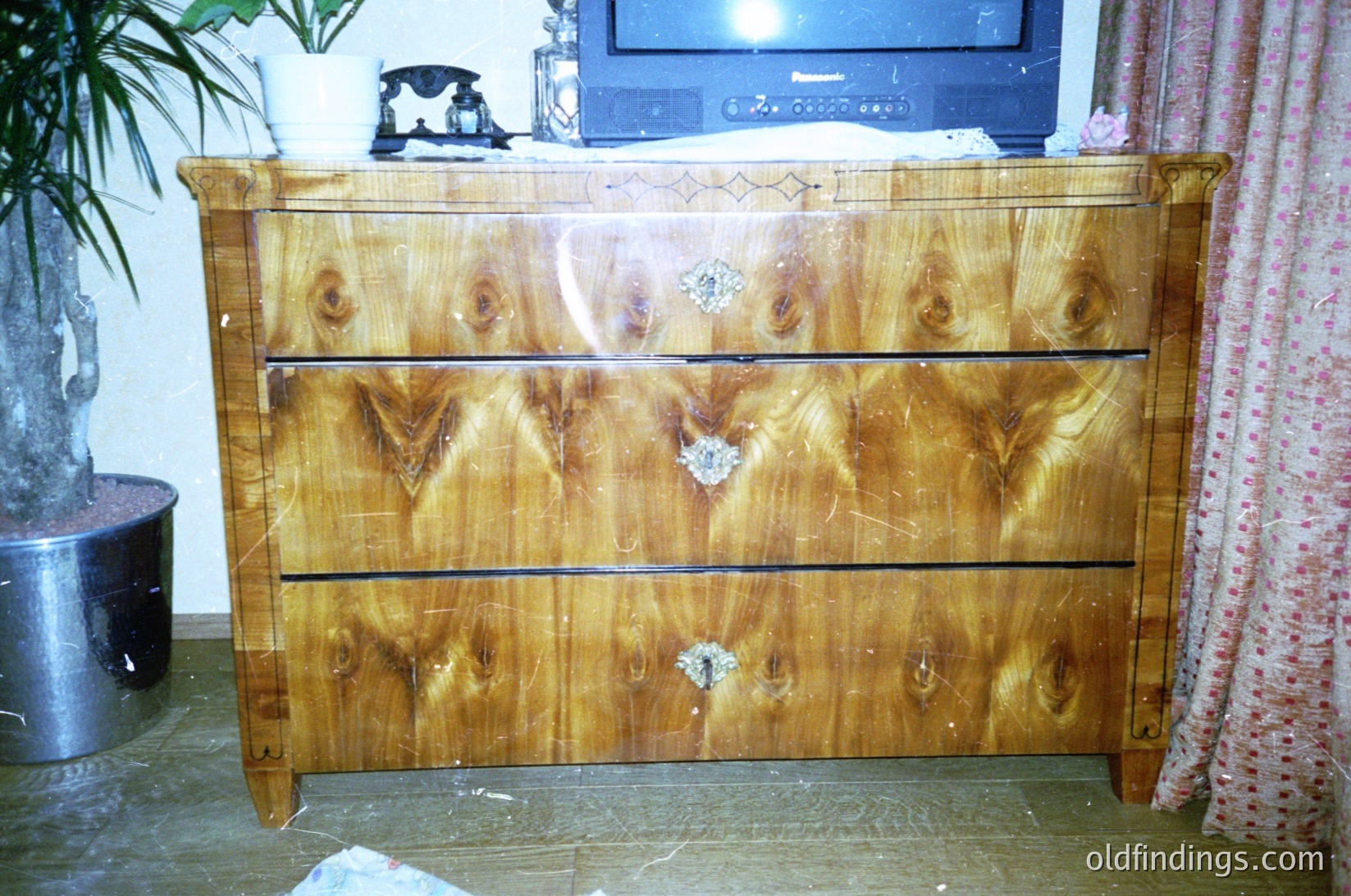 Mid-century modern wooden dresser with three drawers, featuring light oak or birch veneer with visible wood grain and subtle inlays. Slight signs of wear and minor damage (scuffs, dents). Positioned in a domestic interior, likely 1960s–1970s. Background includes a CRT TV, floral-patterned curtain, and indoor plant.