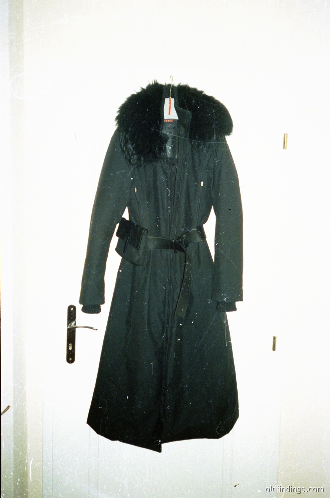Mid-length black wool trench coat with fur-lined hood, belted waist, and visible price tag. Classic 1960s–1970s design, likely European or American. Ideal for fashion archives, vintage clothing research, or historical costume studies.