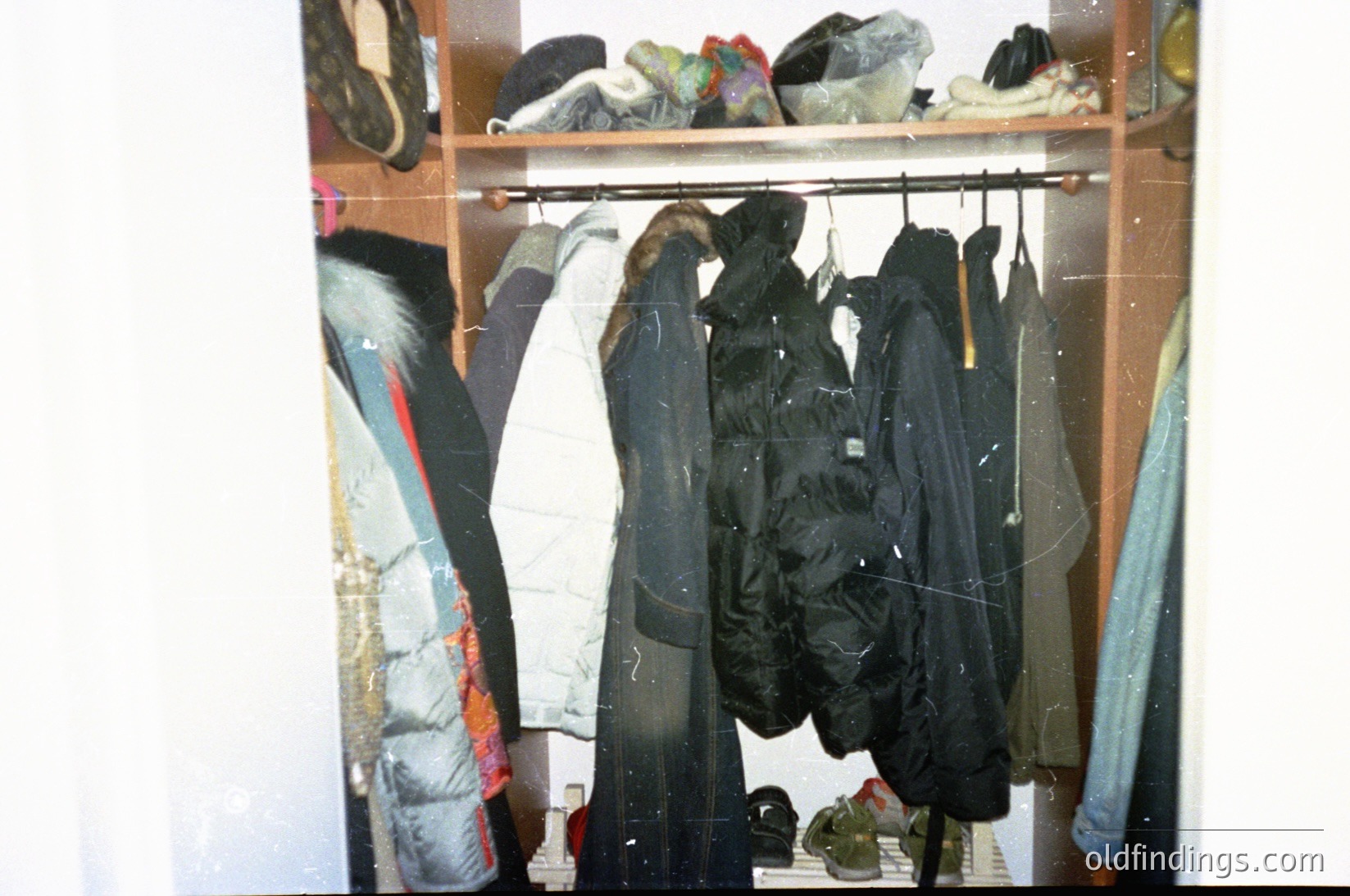 Vintage closet displaying utilitarian outerwear—heavy-duty jackets, raincoats, and insulated parkas hung on a rustic metal rod. Folded items on shelves include boots and folded blankets. Reflective surface suggests aged or low-quality film. Likely mid-20th century domestic setting.
