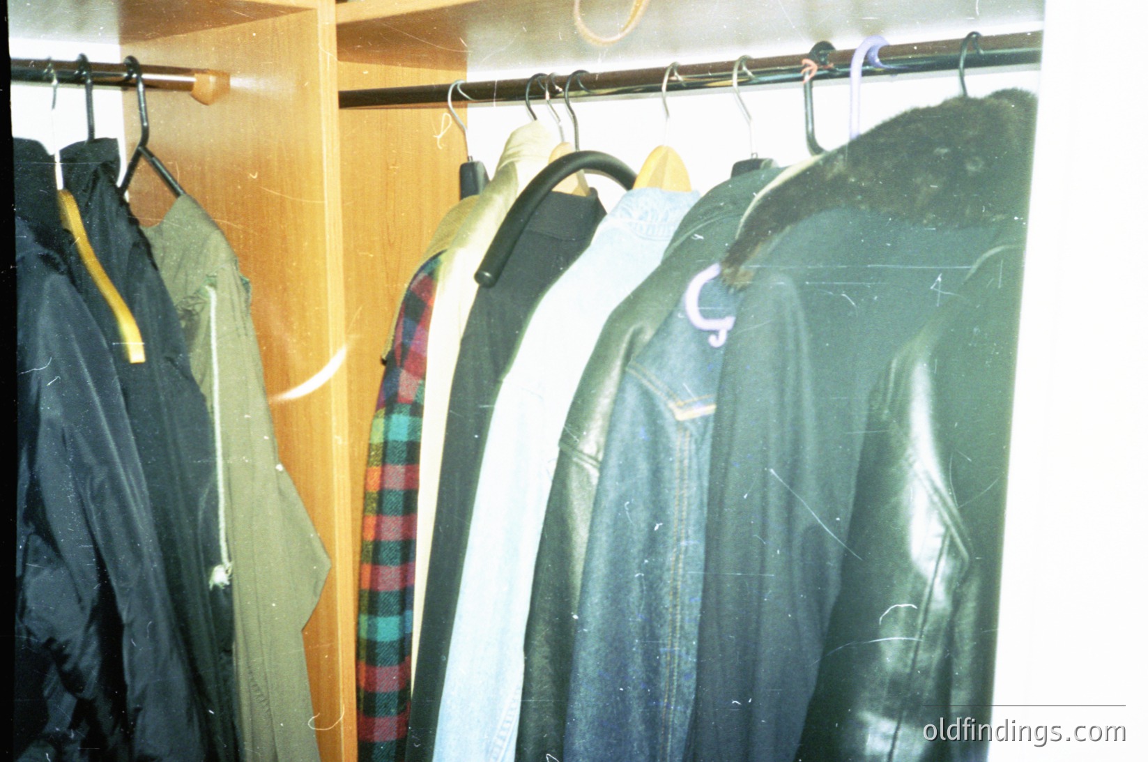 Vintage coat rack featuring 1970s-style outerwear: olive drab utility jacket, green raincoat, plaid windbreaker, and a dark green waxed shell. Hangers on wooden rod against neutral wall. Ideal for historical fashion, military nostalgia, or vintage interior design references.