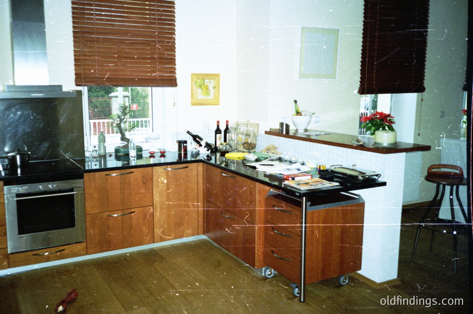 Mid-century modern kitchen with wooden cabinetry and marble countertops, featuring a built-in oven and roll-out prep station. Decor includes framed artwork, potted plants, and a bar stool. Dust and wear suggest an older or abandoned space. Likely or residential interior.