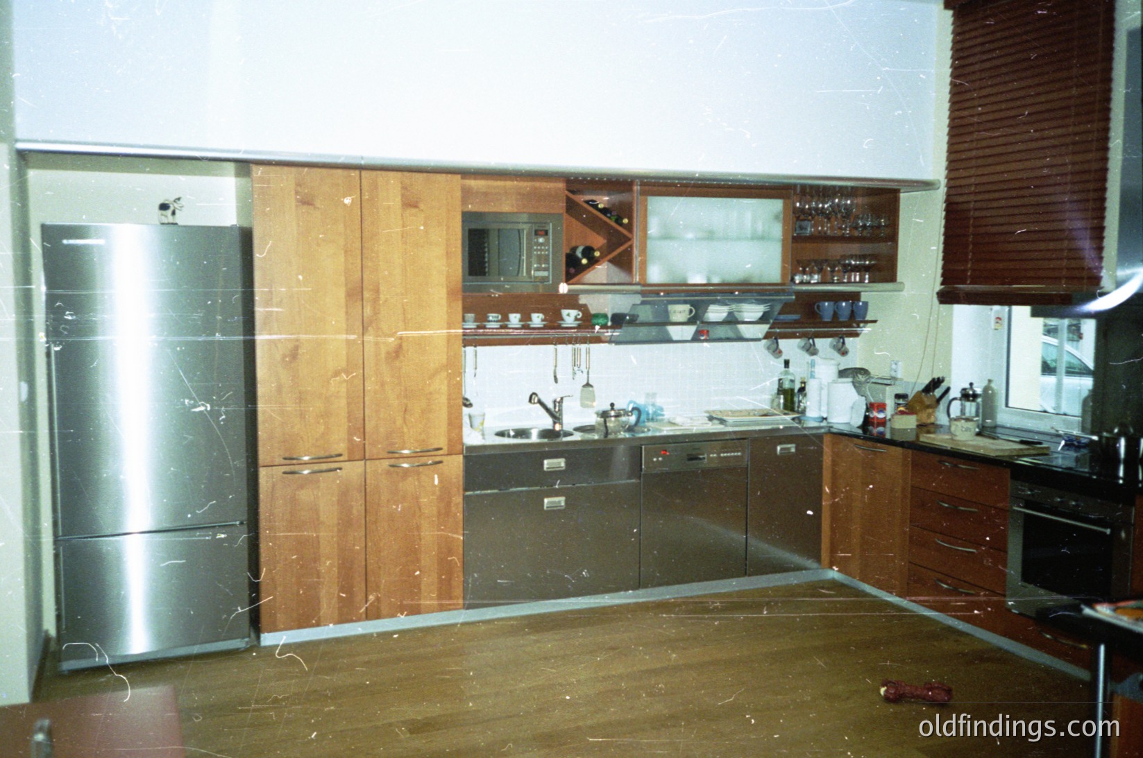Mid-century kitchen with wood-paneled cabinets, stainless steel fridge, and retro appliances. Open shelving holds glassware and dishes. Lighting fixtures and a microwave mounted above the sink. L-shaped layout with linoleum flooring. Likely 1960s–1970s residential interior.