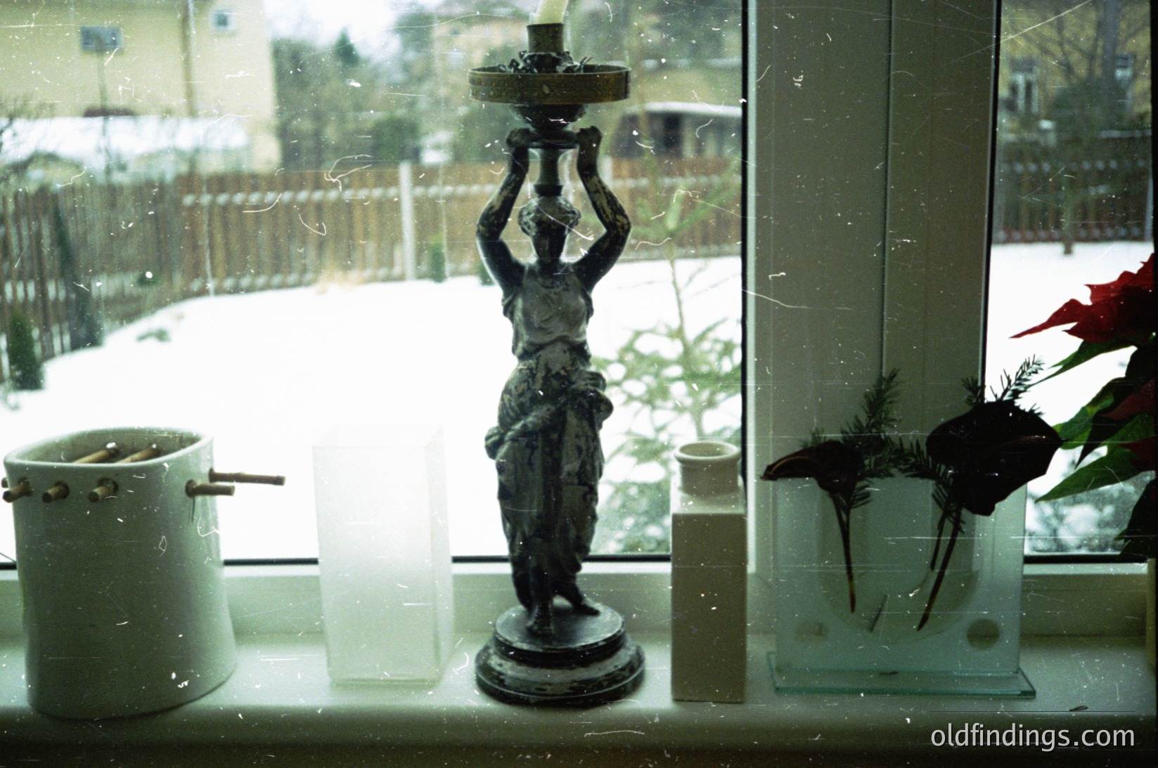 Vintage ceramic lamp with classical female figure holding candle holder, placed on a frosted window sill. Snow-covered garden and residential houses visible outside. White enamel pot and dried floral arrangement in glass vase beside lamp.