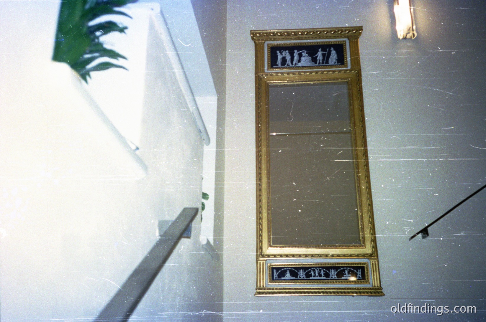 Vintage gold-framed mirror with decorative relief at base, mounted on light marble wall. Reflected image shows a staircase with green foliage. Classic interior design, likely 1950s–1970s.