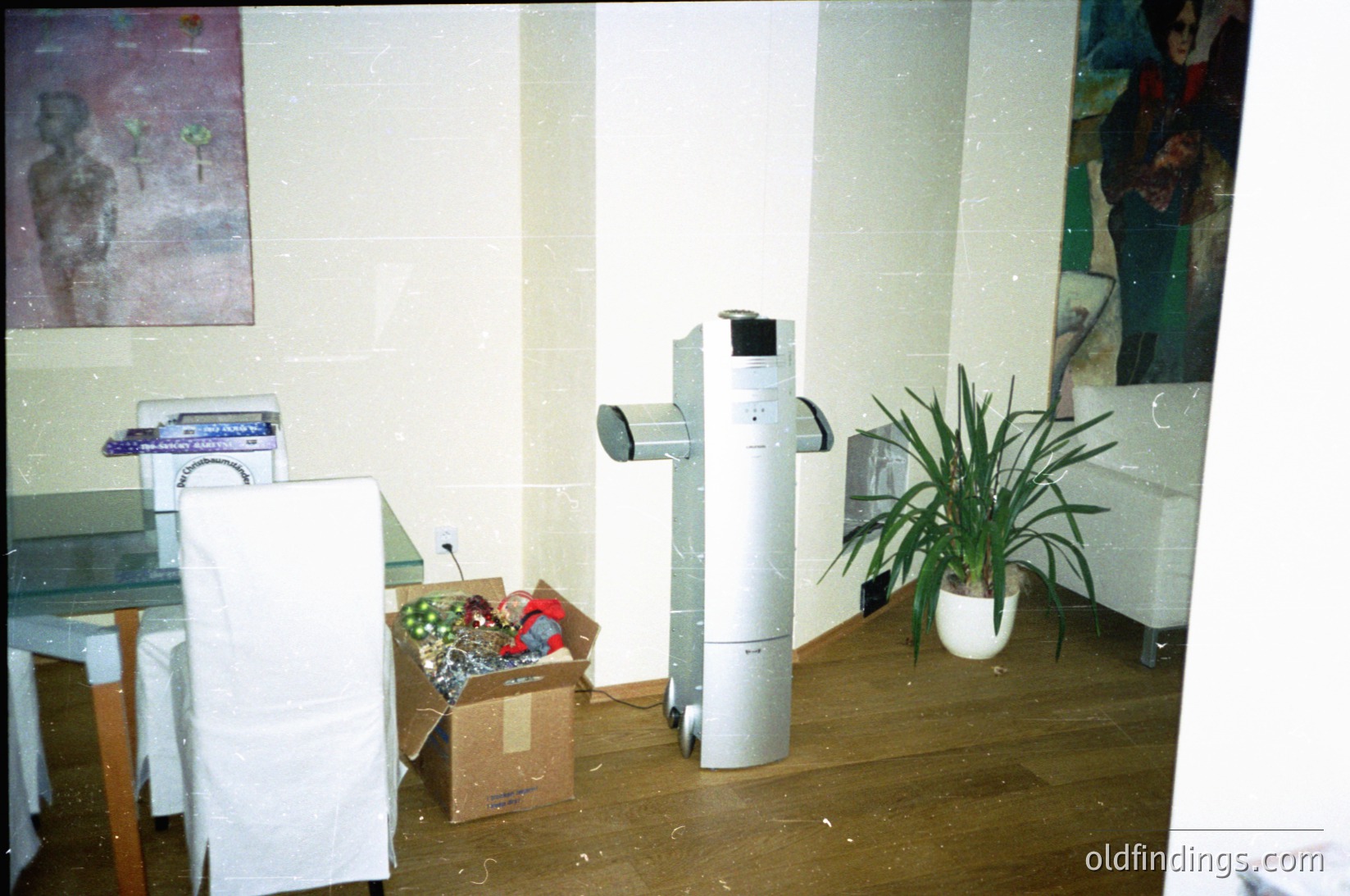 Vintage office space with mid-century design elements: a cylindrical air conditioner unit, a potted plant, and a cardboard box with decorative items. White chairs and tiled walls suggest institutional or commercial use. Likely or era.