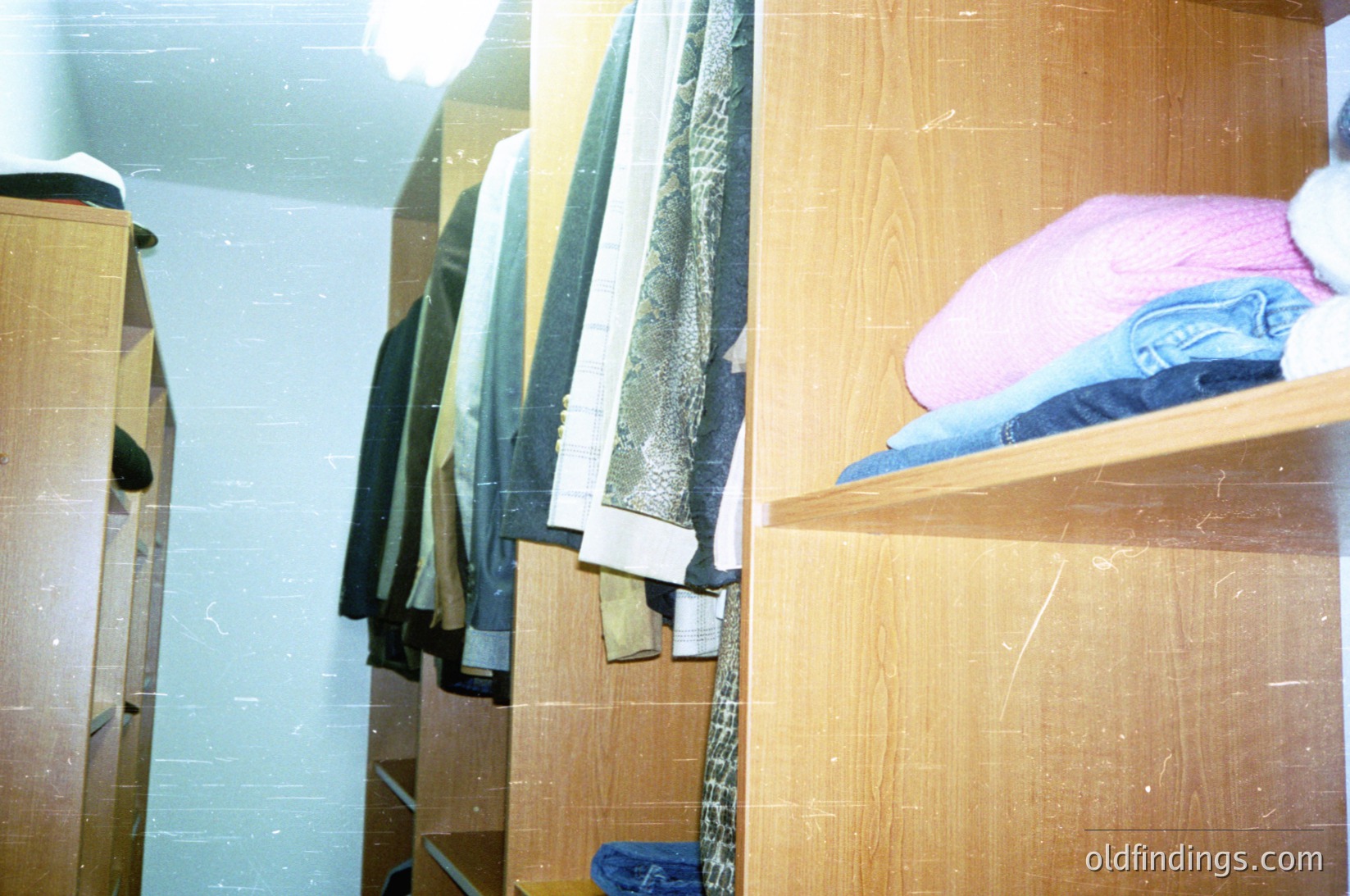 Minimalist wooden closet with vintage storage—hanging coats, folded jeans, and a pink sweater on shelves. Functional, utilitarian design reflects mid-20th-century practicality. Lighting emphasizes worn wood grain and faded fabric textures.