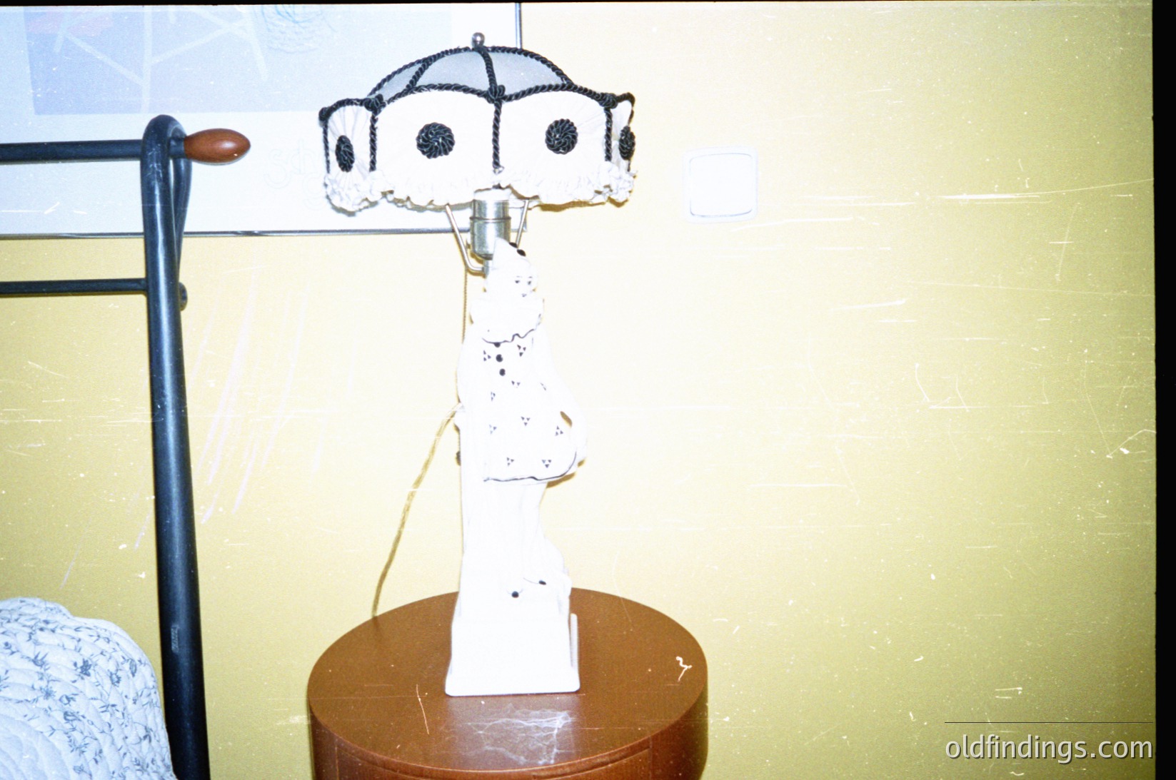 Vintage table lamp with Art Deco-inspired design featuring a sculptural base and black-and-white patterned shade. Placed on a round wooden table beside a metal-framed bed. Mid-century interior with muted yellow walls.