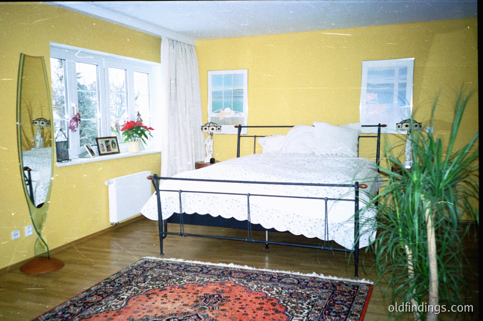 Vintage-style bedroom with two metal-framed beds against yellow walls, separated by a patterned rug. Large windows with white curtains let in natural light, revealing a view of greenery. Decor includes a potted plant, framed photos, and a radiator. Likely Eastern European mid-century design.