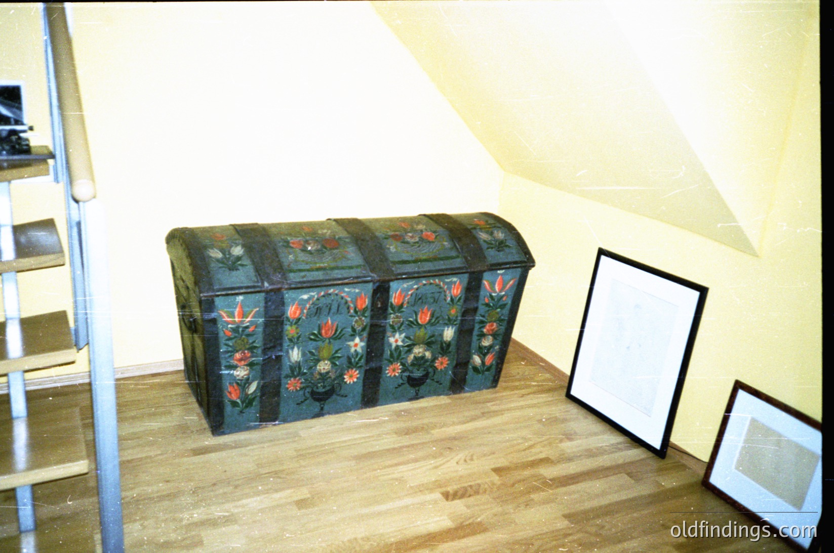 Vintage wooden trunk with floral motifs in deep blues, reds, and golds, placed on wooden flooring. Empty frames and metal stair railing visible. Likely Eastern European folk art, 19th–early 20th century.