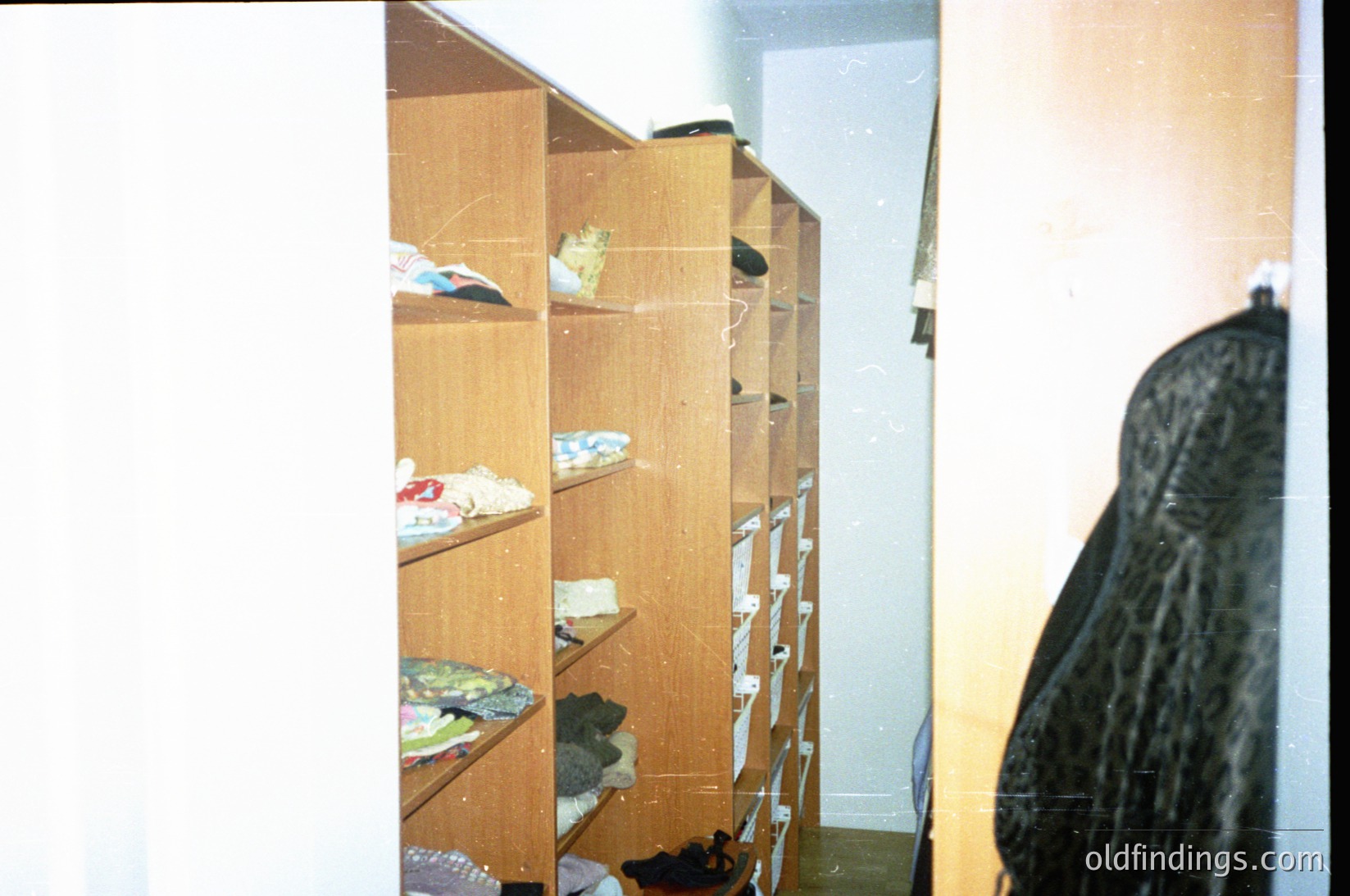Vintage wooden closet with open shelves holding folded clothes, shoes, and plastic-wrapped items. Lighting suggests indoor residential setting, likely mid-20th century.