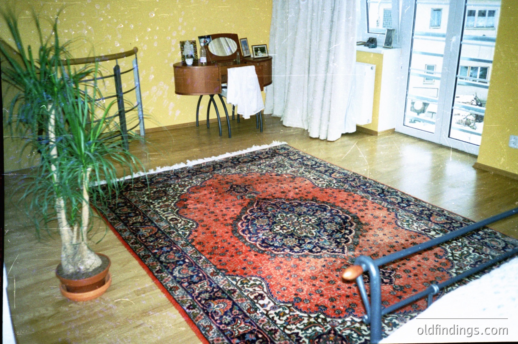 Mid-century modern bedroom with vintage furnishings: ornate red/black patterned rug, wooden vanity with mirror, and potted plant. Balcony railing and cityscape view through large windows. Yellow wallpaper and white curtains add retro charm. Likely 1960s–1970s European interior.