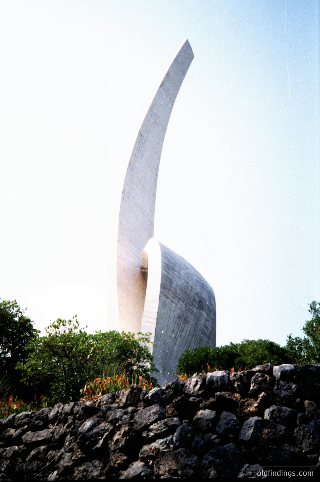 Modernist concrete sculpture resembling a stylized bird or abstract wing, mounted on a stone base. Likely part of a public art installation or urban landscape design. Distinctive curved form and minimalist aesthetic.