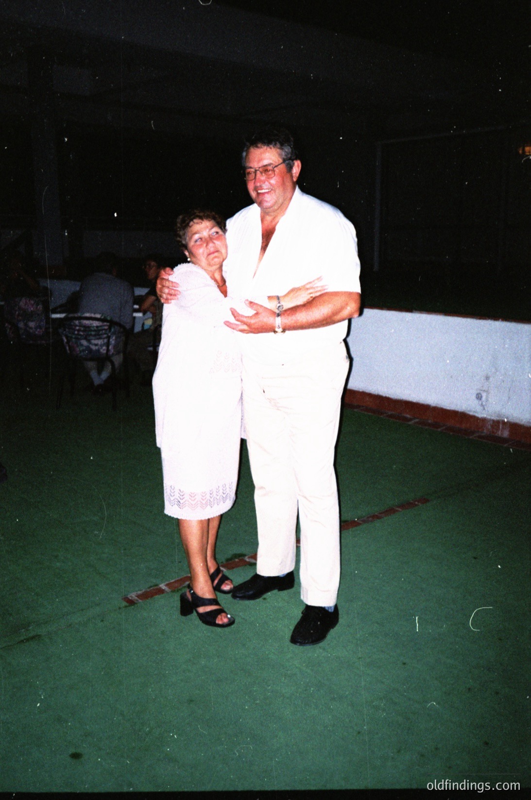 Couple dancing in matching white attire at an indoor event, likely a 1980s–1990s social gathering. Woman wears a white lace dress with sandals; man in white pants and shirt with a watch. Green vinyl dance floor and blurred background suggest a community hall or recreational center.