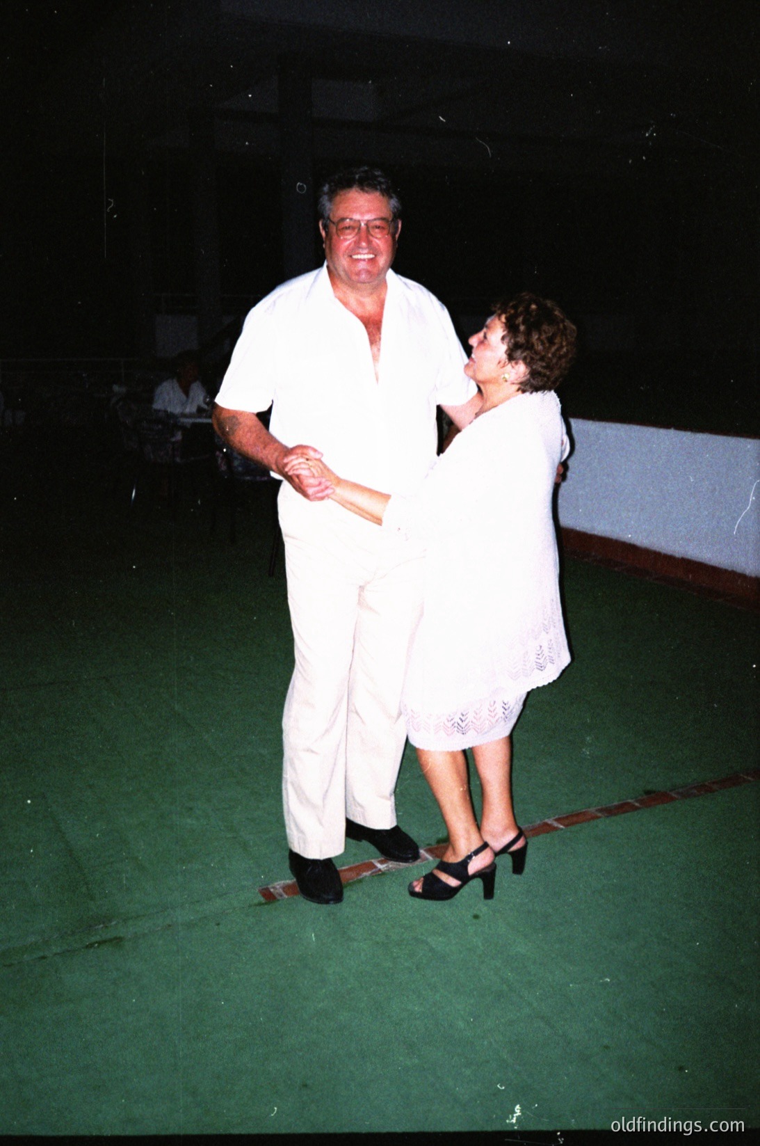 Couple dancing in a dimly lit indoor venue, likely a 1980s/1990s social event. Man in white suit, woman in sleeveless white dress with lace trim. Green dance floor with blurred background suggesting music or crowd.