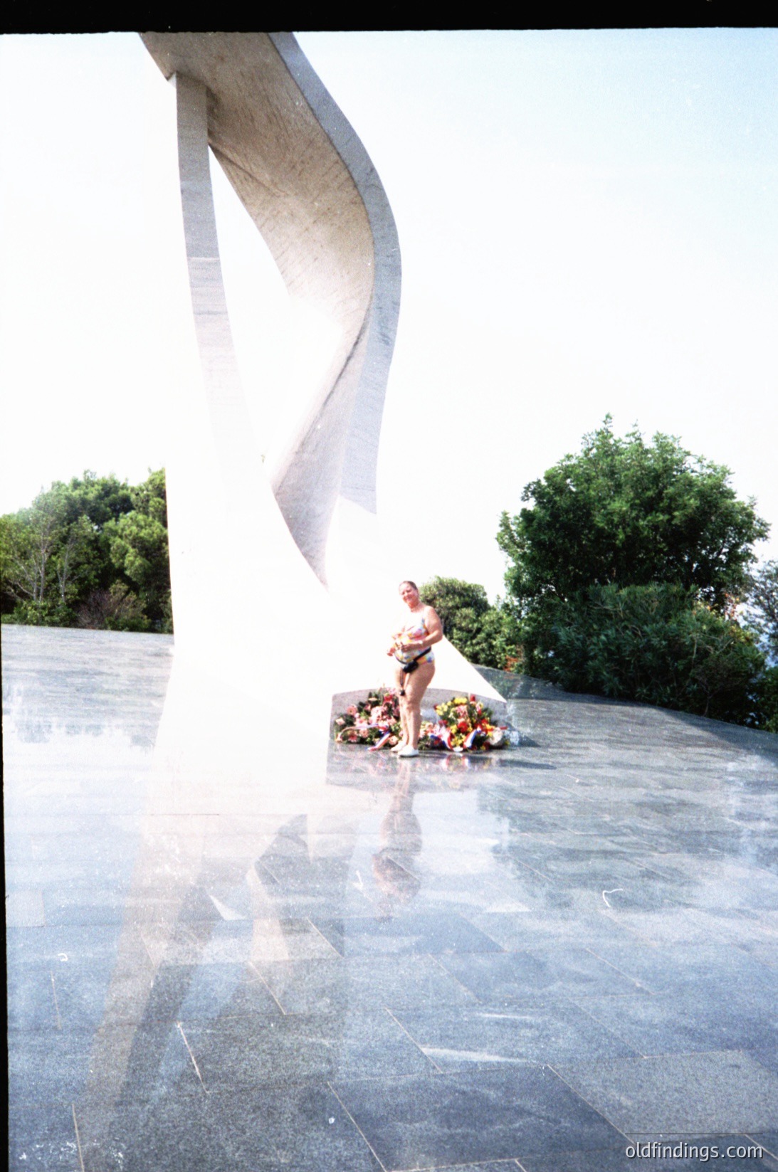 A solemn individual kneels in quiet reflection at a war memorial, placing flowers atop a wreath. The modernist concrete monument features sweeping, abstract curves. Sunlight casts sharp shadows on the polished stone plaza. Likely Eastern European, 1980s–1990s era.
