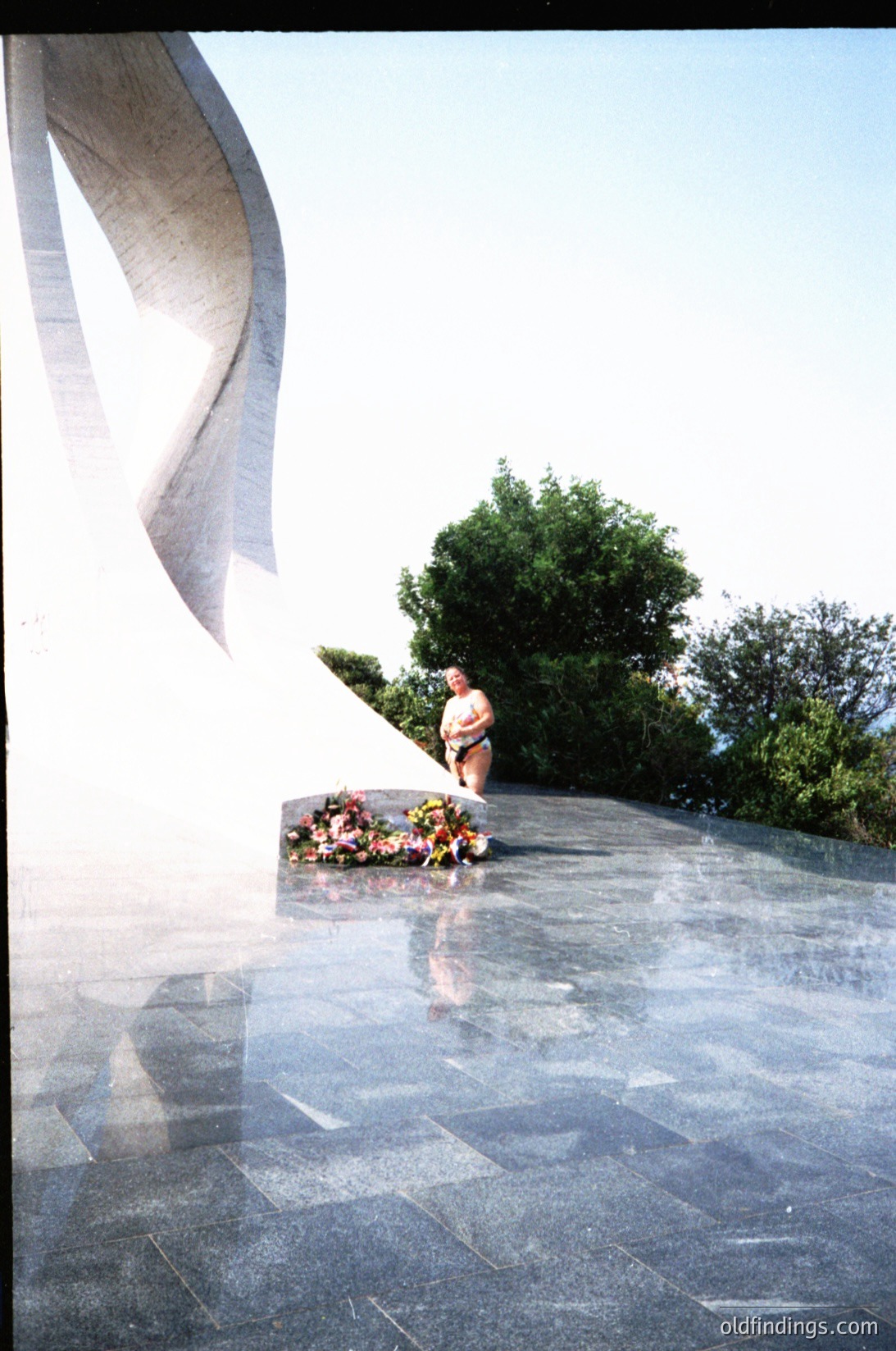 Modernist concrete monument with sweeping, wave-like design. A person kneels beside floral tribute on reflective stone plaza. Lush greenery and trees frame the scene. Likely a memorial or tribute site.