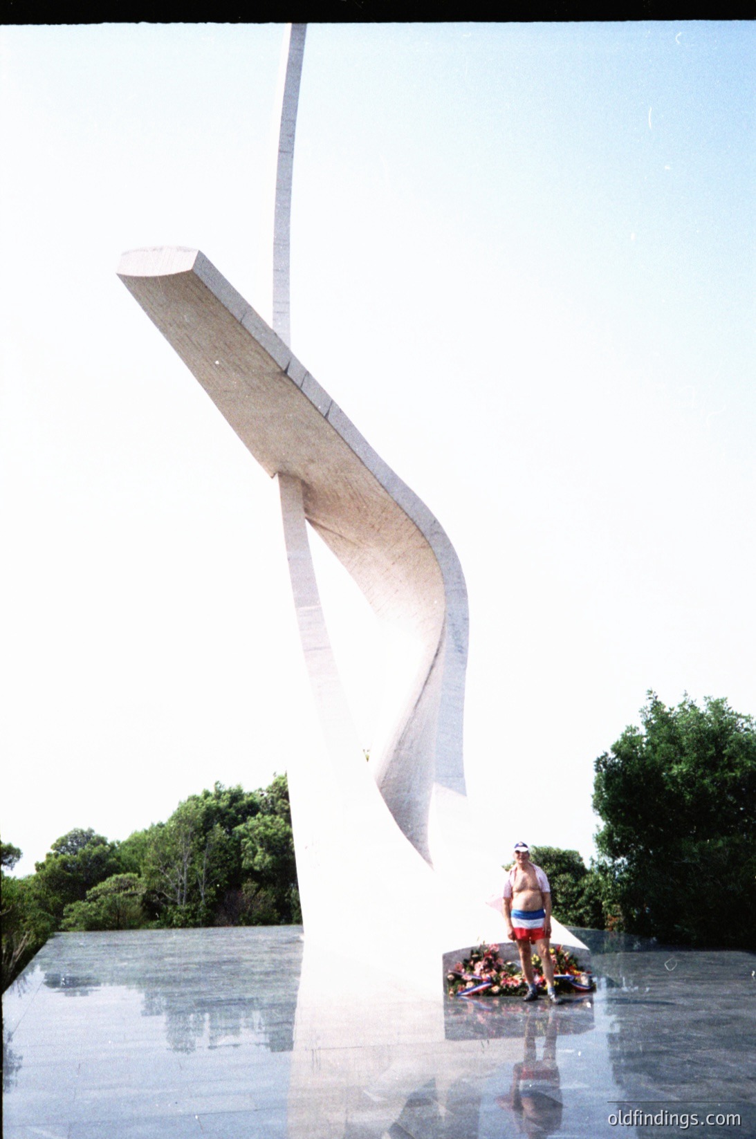 Modernist concrete sculpture resembling a wave or sail, anchored in reflective water. A person in swimwear poses beside it, suggesting a public park or memorial setting. Likely mid-20th century (1950s–1970s) based on style.