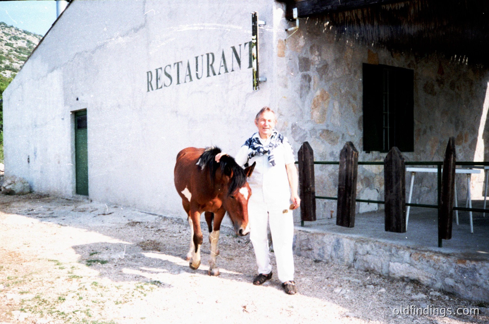 Rustic stone building with "RESTAURANT" sign in bold letters, featuring a man in light-colored attire leading a brown horse near a wooden fence. Likely alpine or rural European setting, mid-20th century.