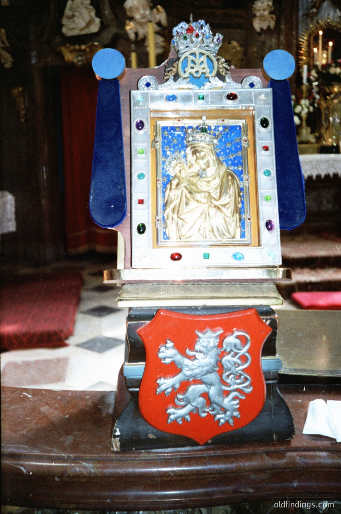 Religious icon housed in an ornate 19th-century Czech shrine, featuring a crowned Madonna and Child framed by blue velvet drapes and a red shield with a crowned lion emblem. Intricate mosaic and gemstone details enhance the wooden base. Likely from a historic Orthodox or Catholic church.