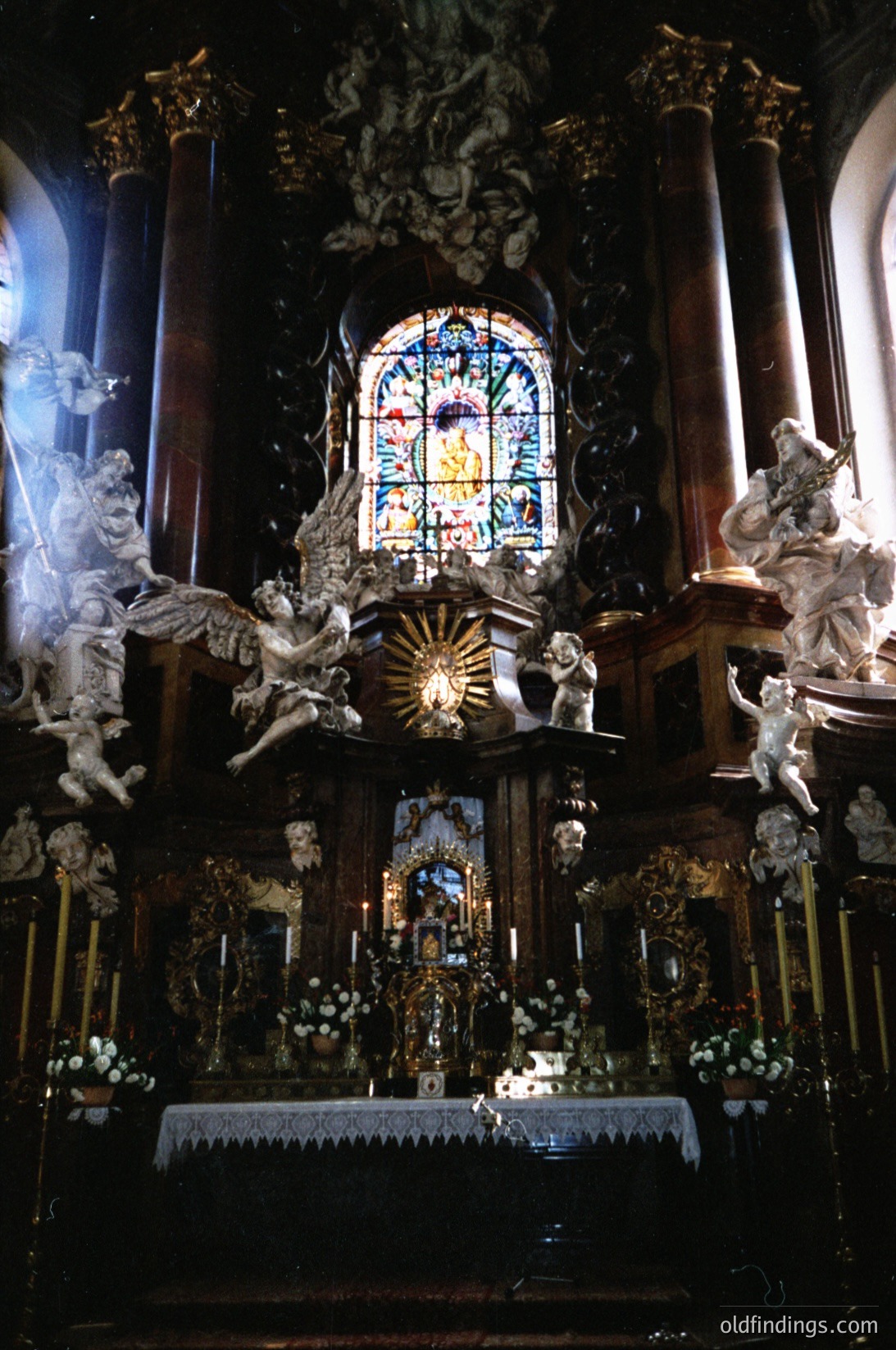 Baroque-style altar featuring gilded woodwork, intricate carvings, and a central stained-glass window depicting religious iconography. Flanking angels and sculpted cherubs enhance the ornate design, with floral arrangements and candle holders adding detail. Likely European, 17th–18th century.