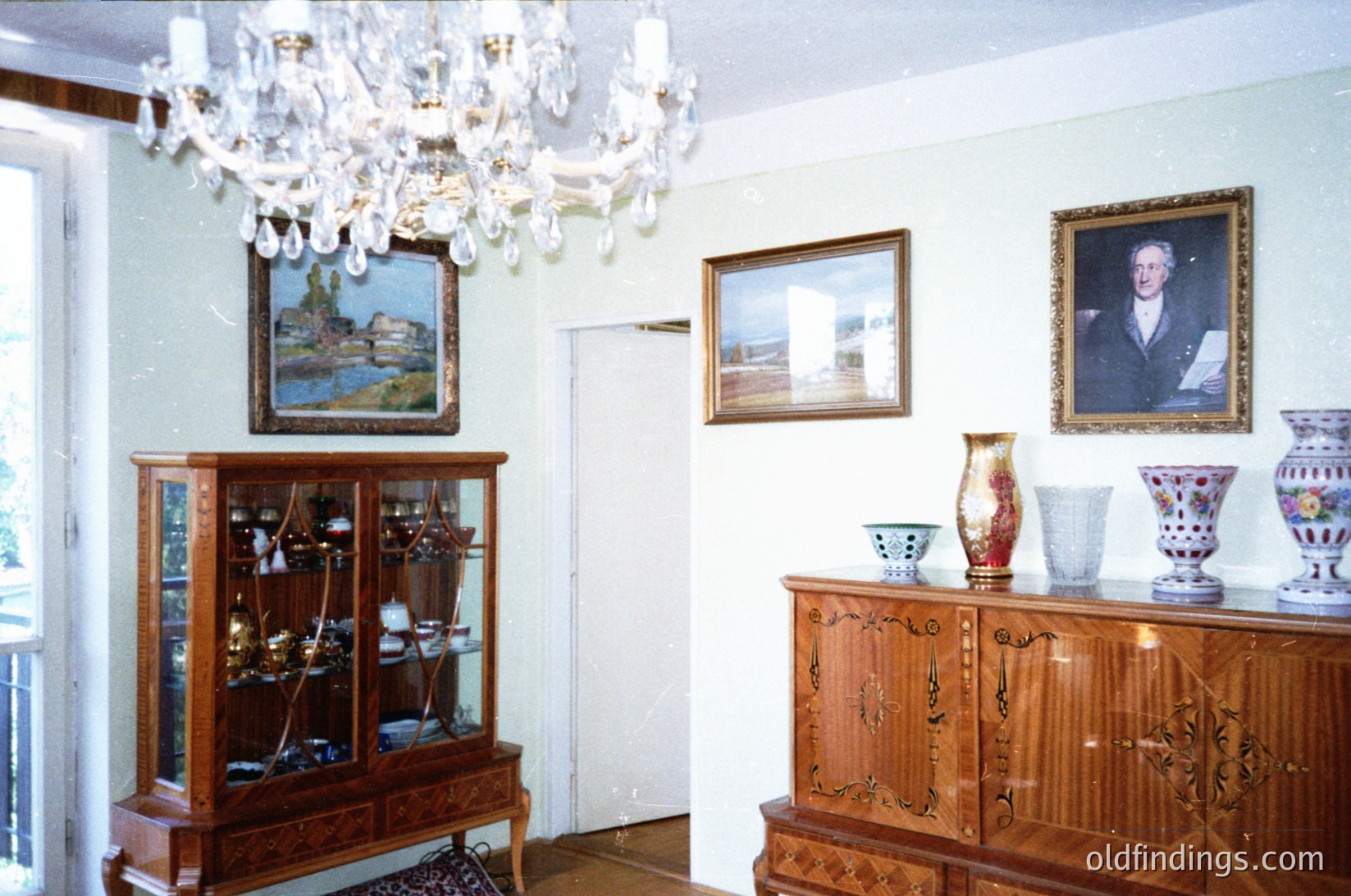 Vintage interior featuring ornate wooden furniture, including a glass-fronted cabinet displaying silverware and a carved sideboard with decorative vases. Three framed portraits and a landscape painting adorn the walls under a crystal chandelier. Lighting suggests a mid-20th-century European home, likely or .