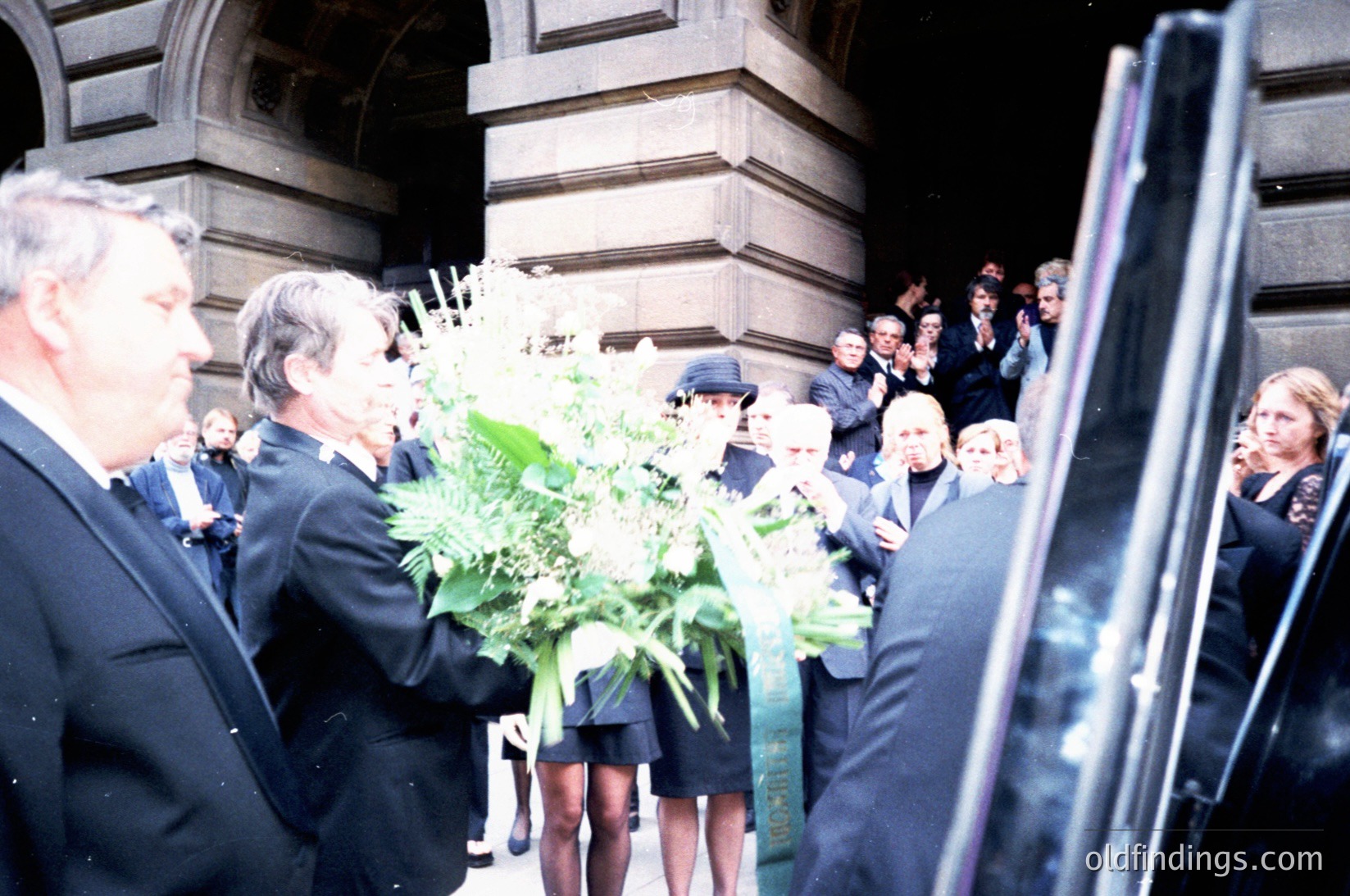 Formal gathering at an ornate stone building entrance, likely a funeral or memorial. Attendees in 1970s-era suits and dresses hold large floral wreaths. Crowd stands on steps, some in profile, others facing the camera. Classic architecture with arched windows and detailed stonework visible.